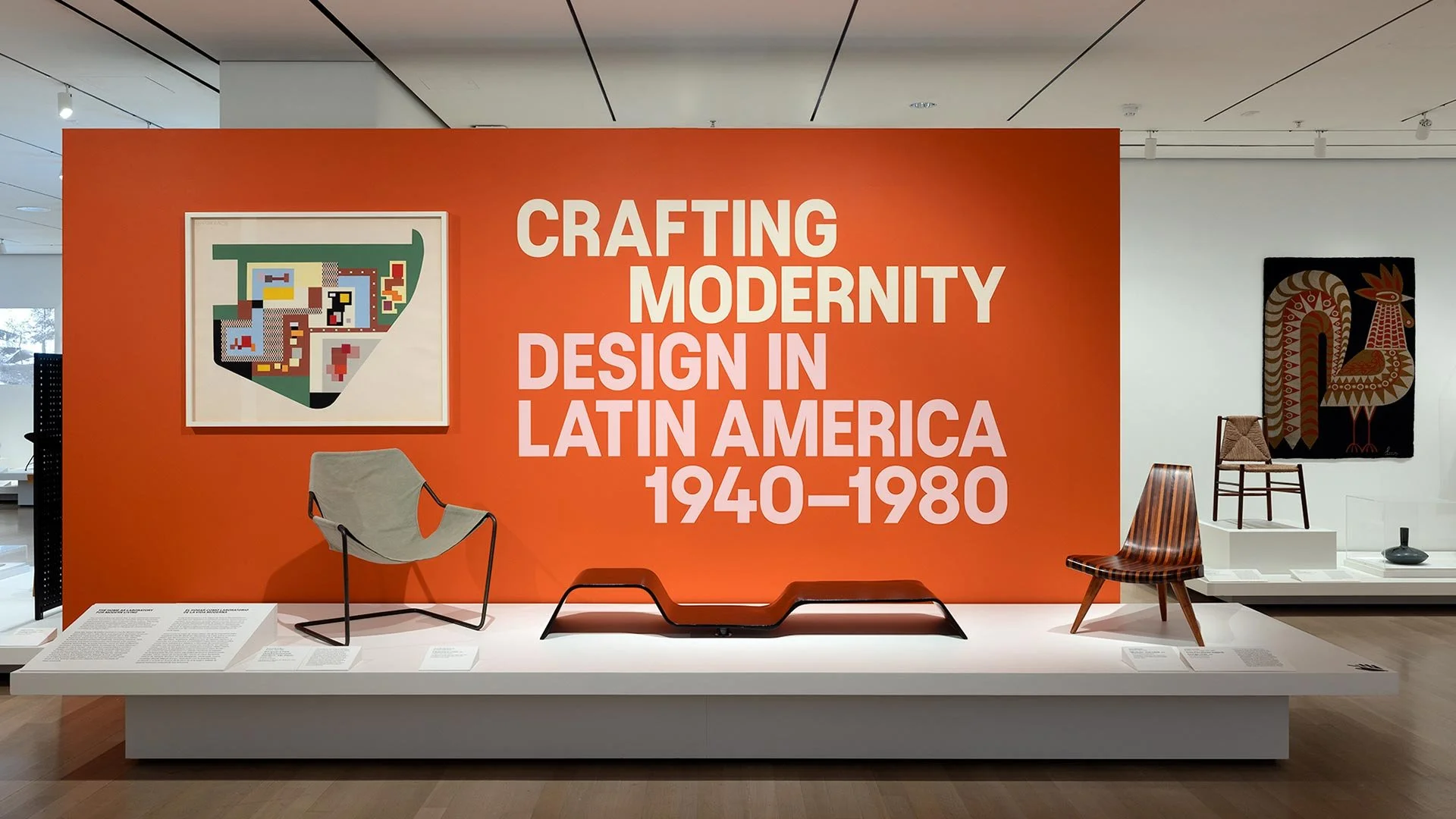 Exhibition display with the title 'Crafting Modernity: Design in Latin America 1940-1980', featuring modern furniture and artwork, including a framed pixel art piece and textile art.