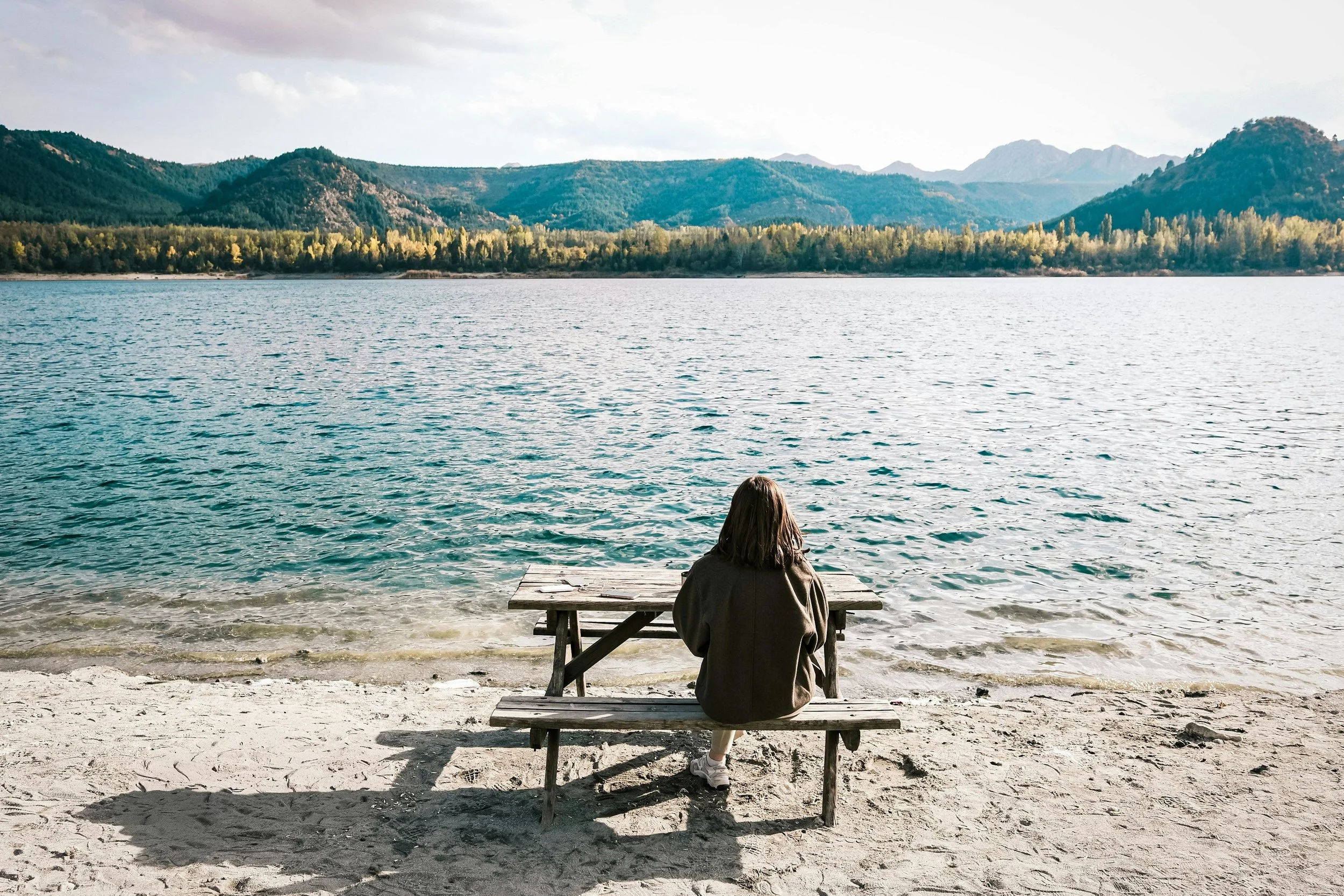 Person sitting on a wooden bench by a lake, facing the water with mountains in the background.