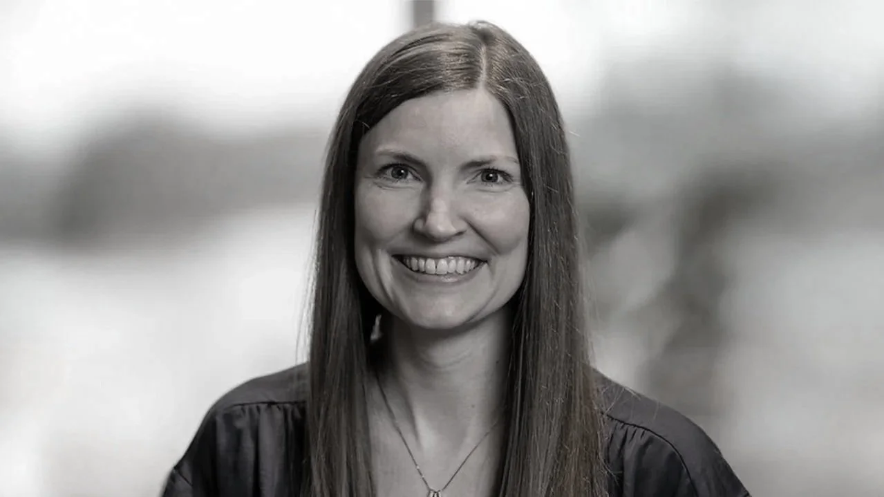 A black-and-white portrait of a smiling woman with long, straight hair, wearing a dark top.