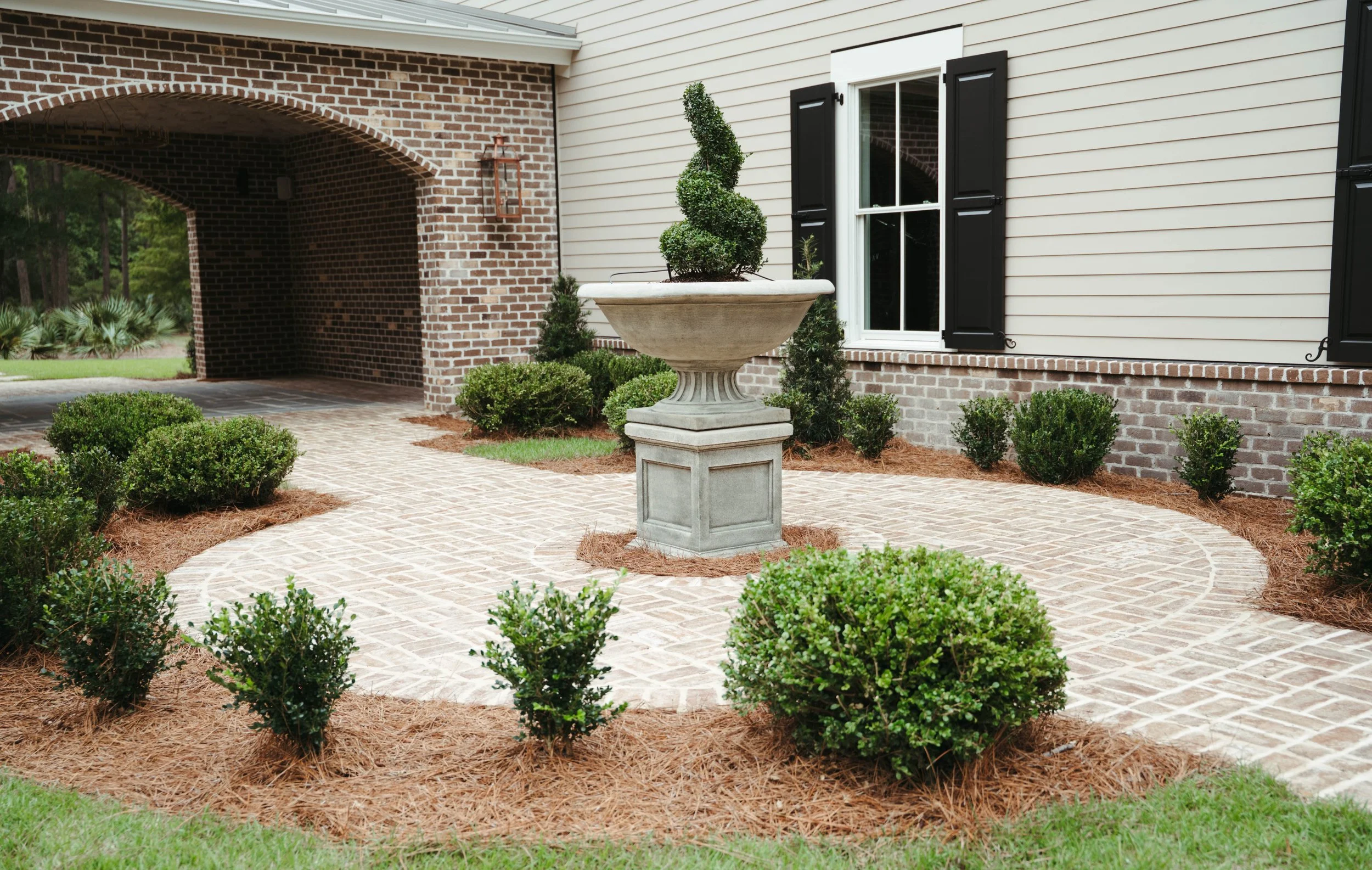 A front yard with a brick and siding house, neatly trimmed bushes, and a stone statue on a brick pathway.