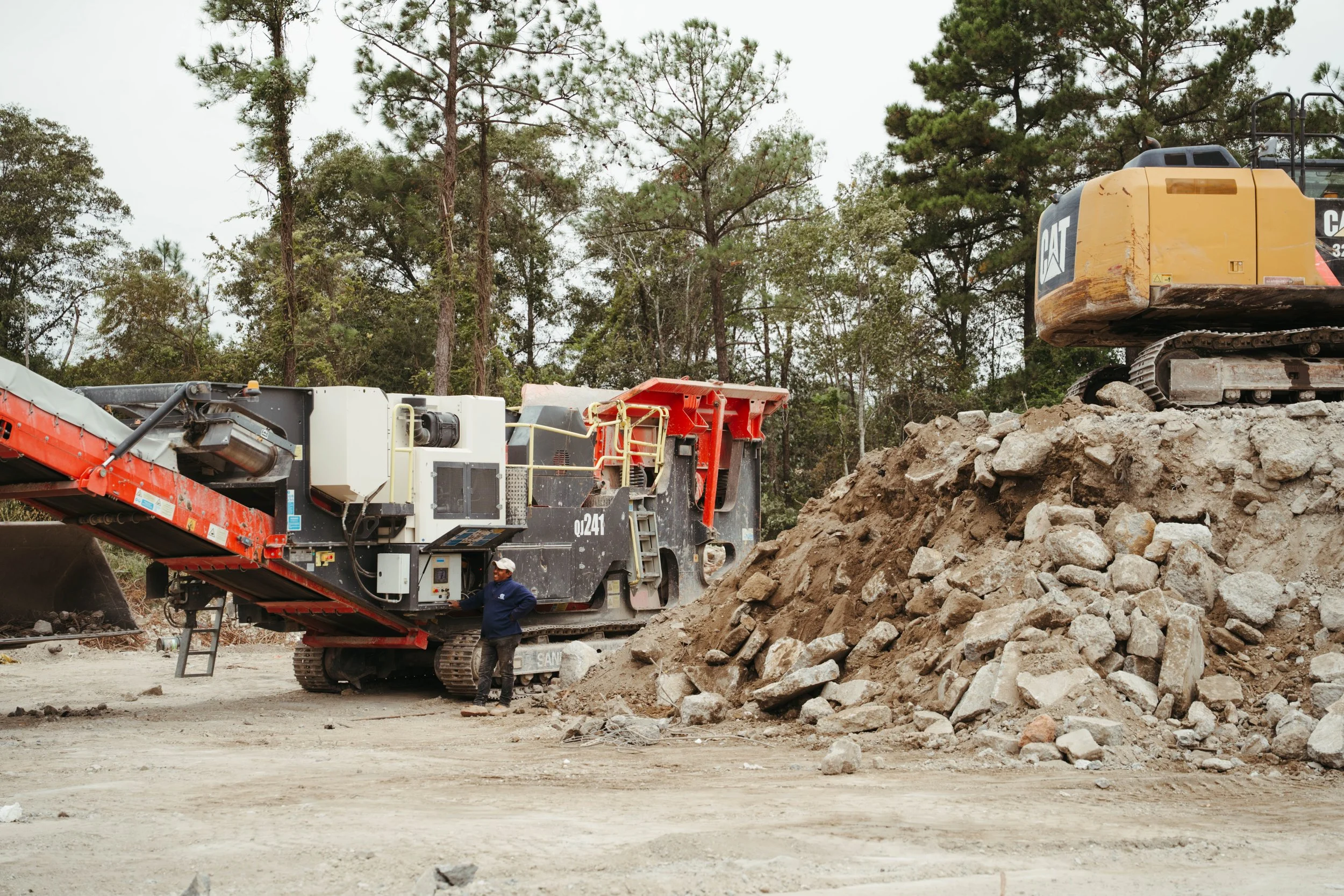 Worker standing beside construction machinery and a pile of dirt and rocks at a construction site surrounded by trees.