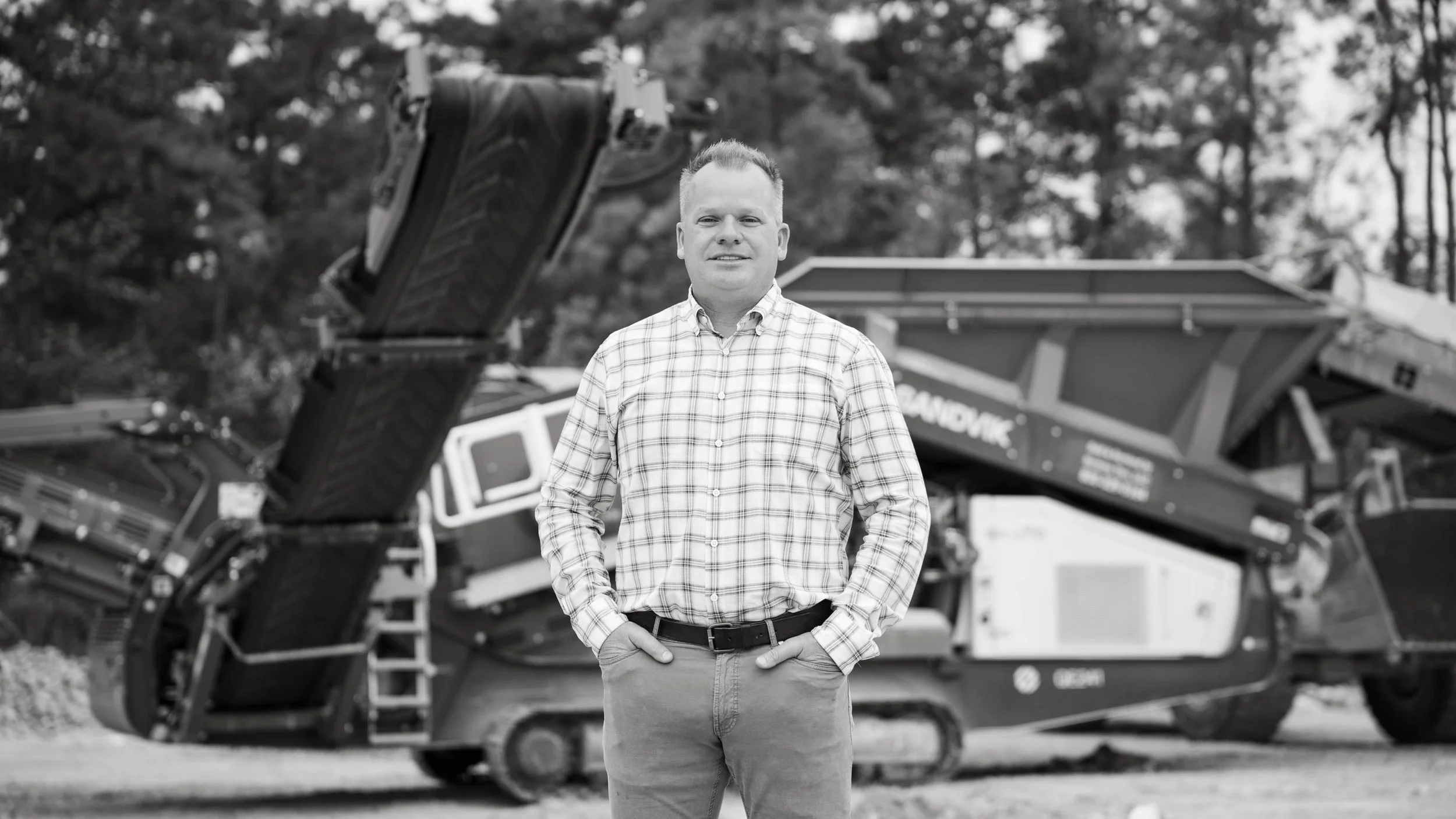 A man in a plaid shirt and khaki pants standing outdoors with a large piece of construction equipment in the background.