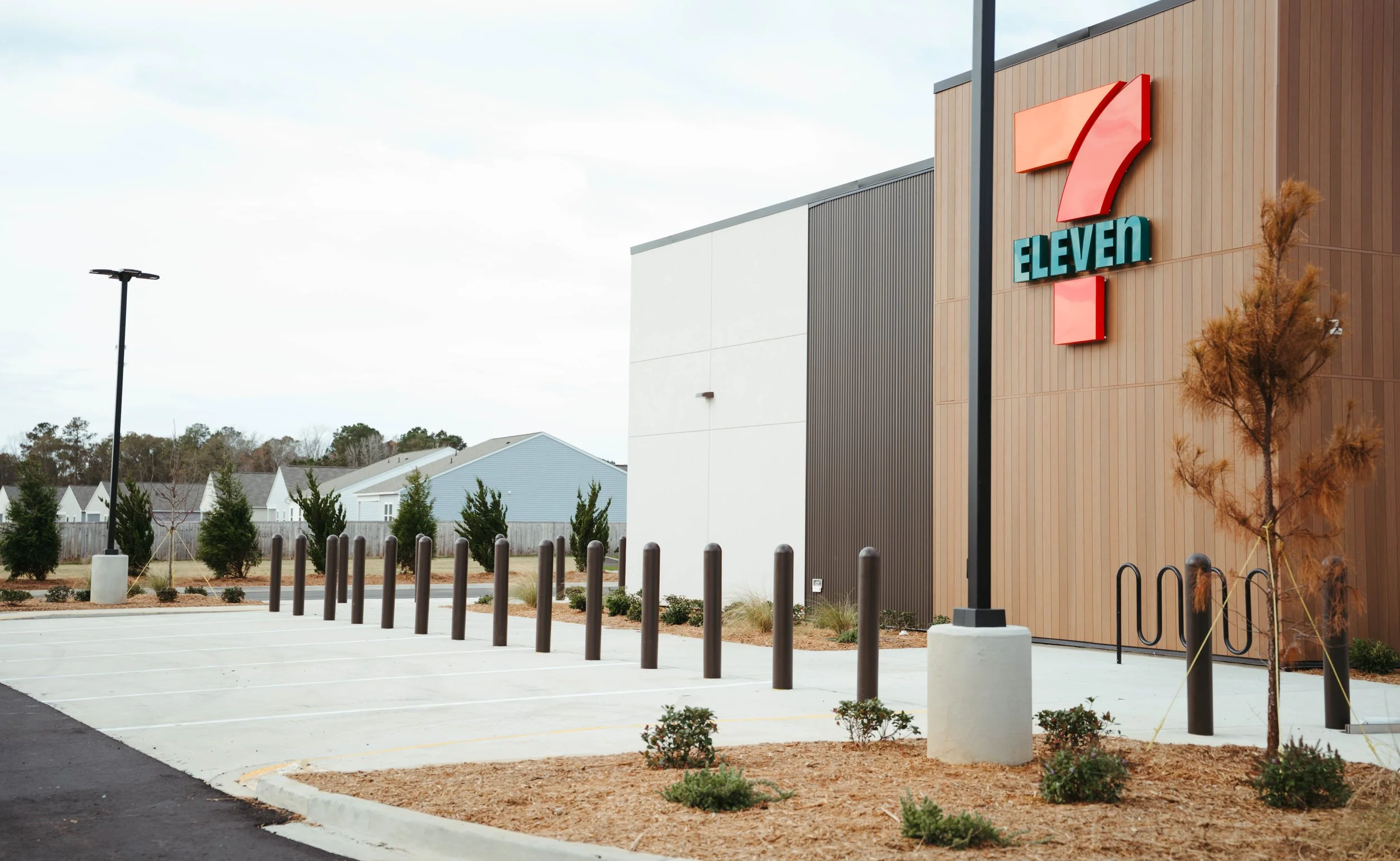 An empty parking lot in front of a 7 Eleven convenience store with some small trees and bushes, a lamppost, and the store's sign on the building with a wood and white exterior.