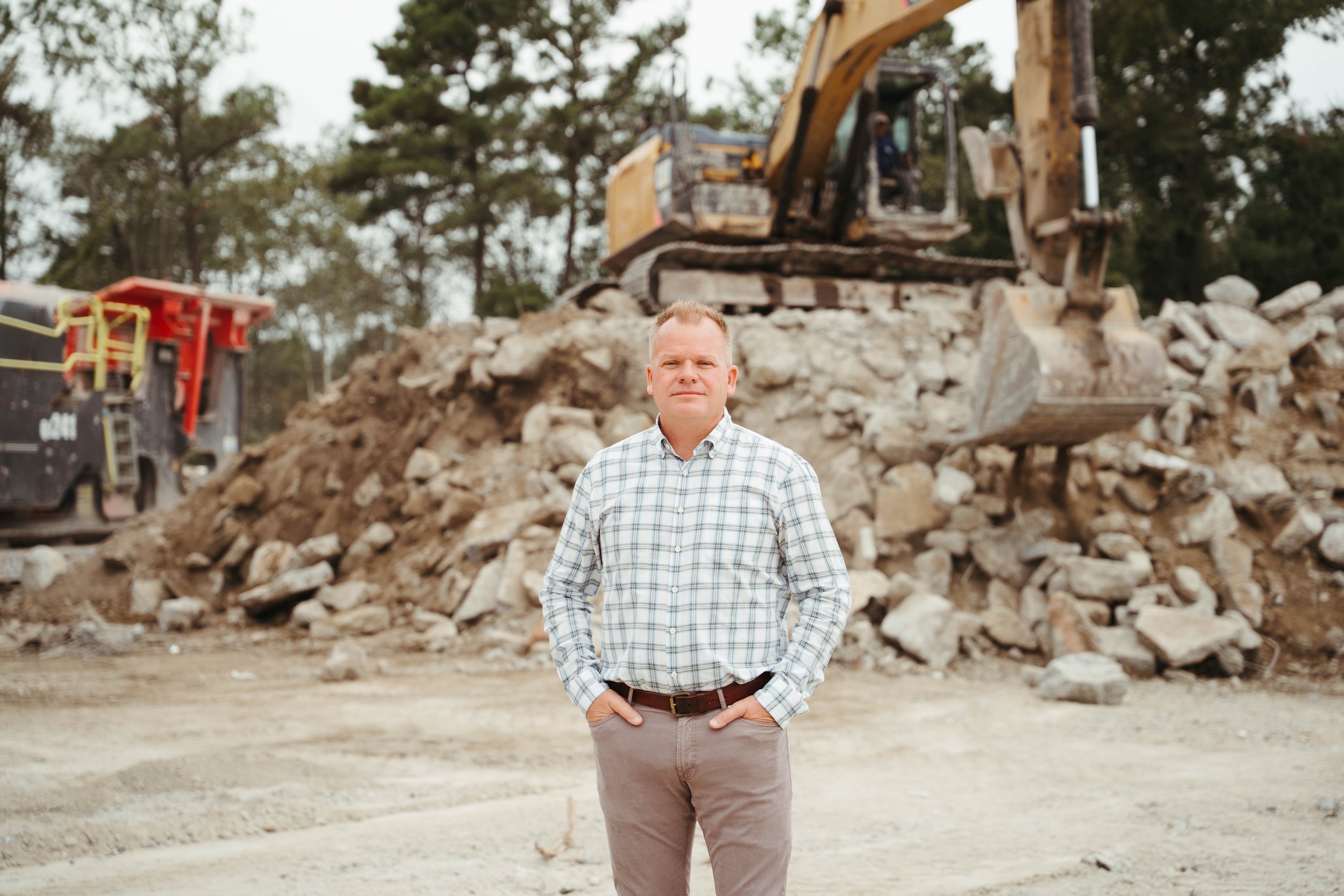 A man wearing a plaid shirt and gray pants stands with hands in pockets in front of a construction site with an excavator and pile of rubble and rocks.