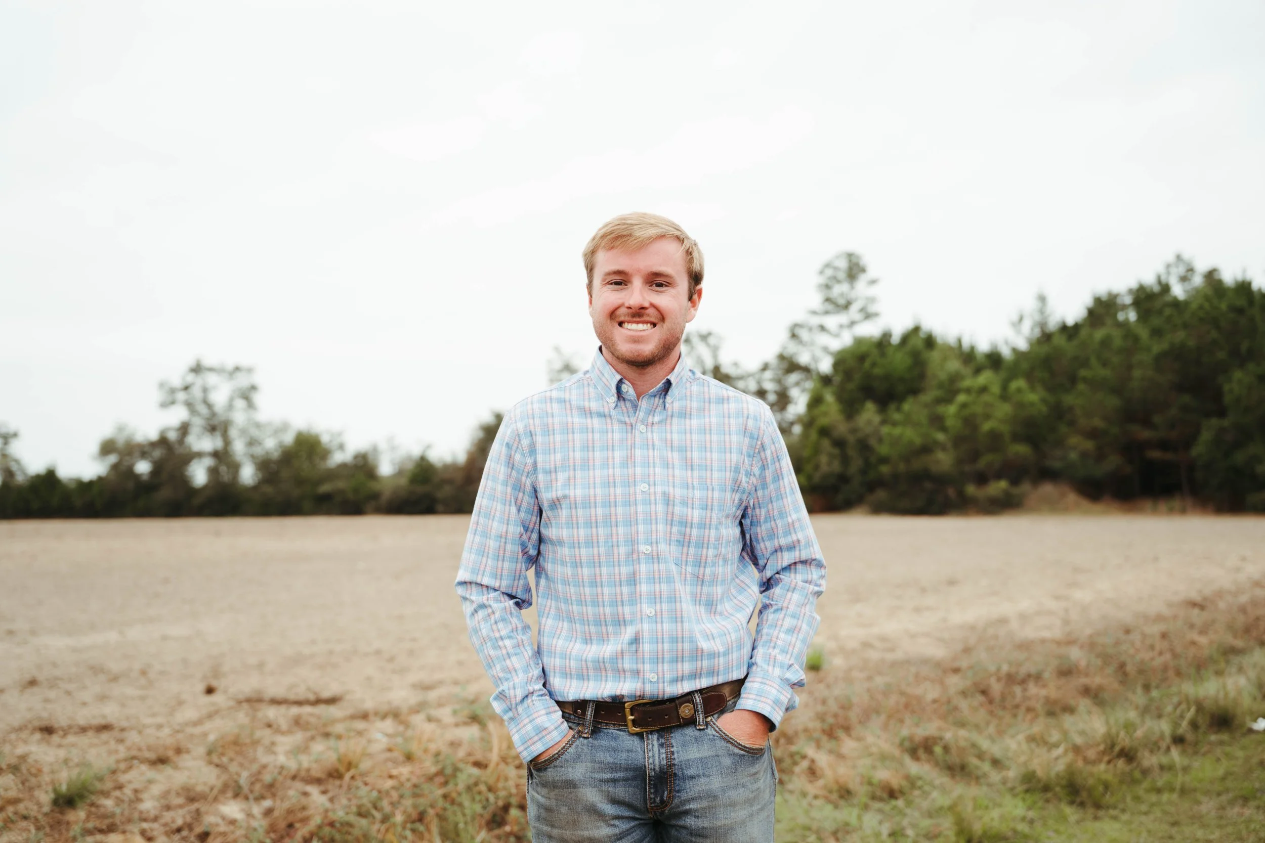 A young man with blonde hair and a light beard standing outdoors in a field, smiling at the camera, wearing a plaid shirt and blue jeans.