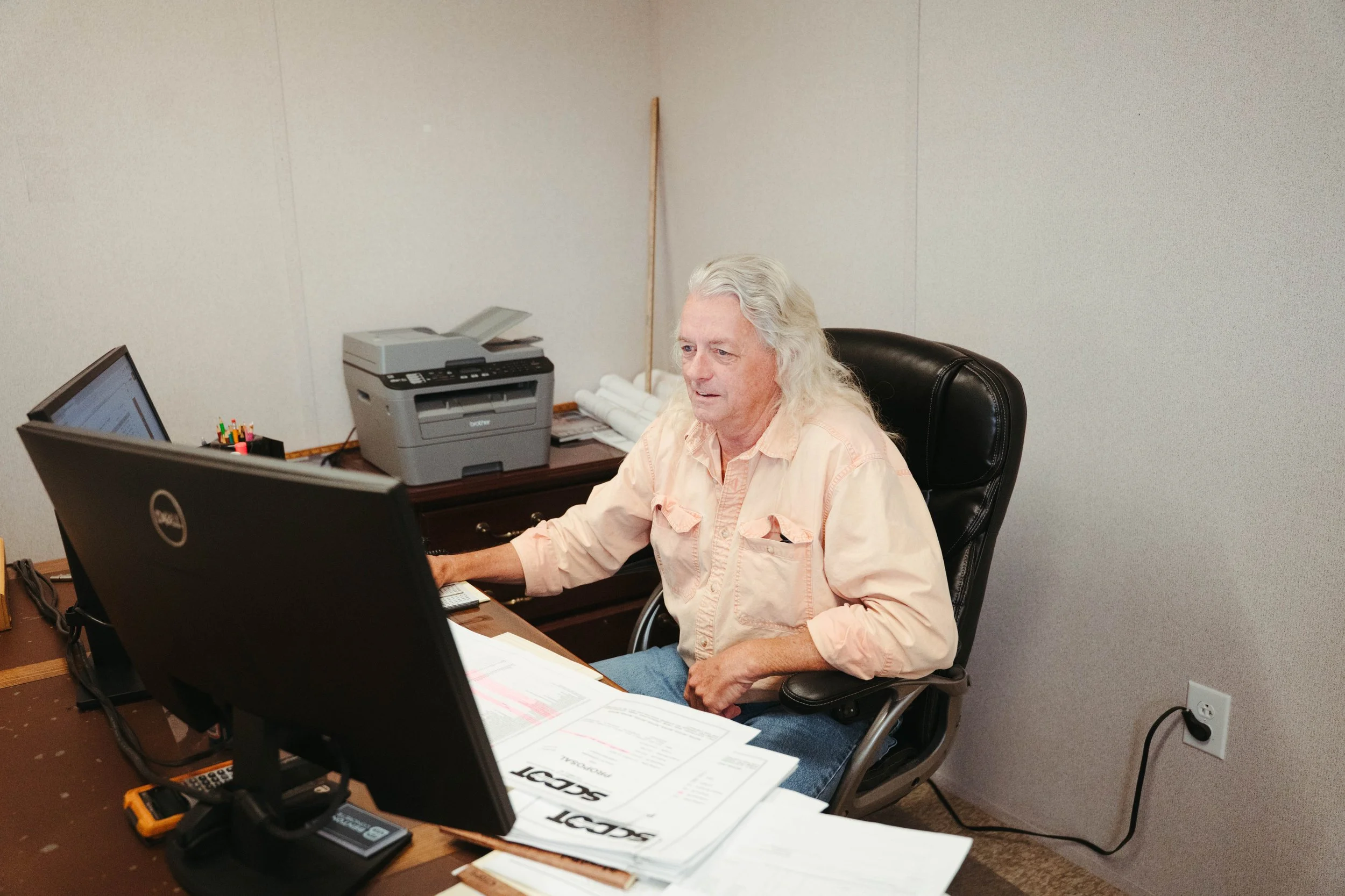 An elderly man with long gray hair wearing a peach shirt working at a cluttered desk with a monitor, papers, pens, a printer, and a cordless phone.