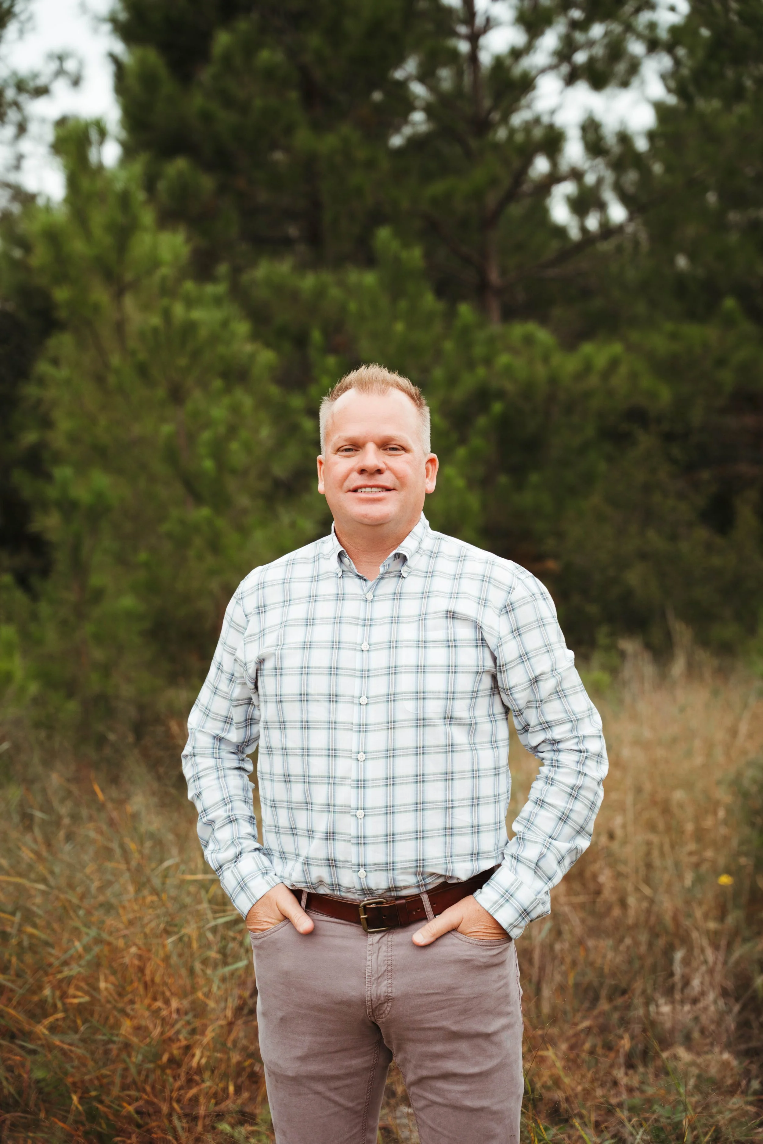 A man standing outdoors in front of green trees, wearing a plaid shirt and khaki pants, with hands in pockets, smiling at the camera.
