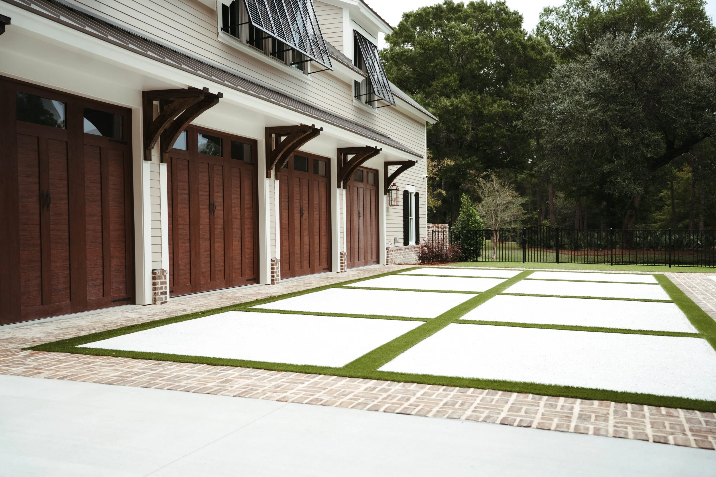 Residential garage with four wooden doors, white siding, brick accents, and a concrete and brick driveway, with green grass and trees in the background.