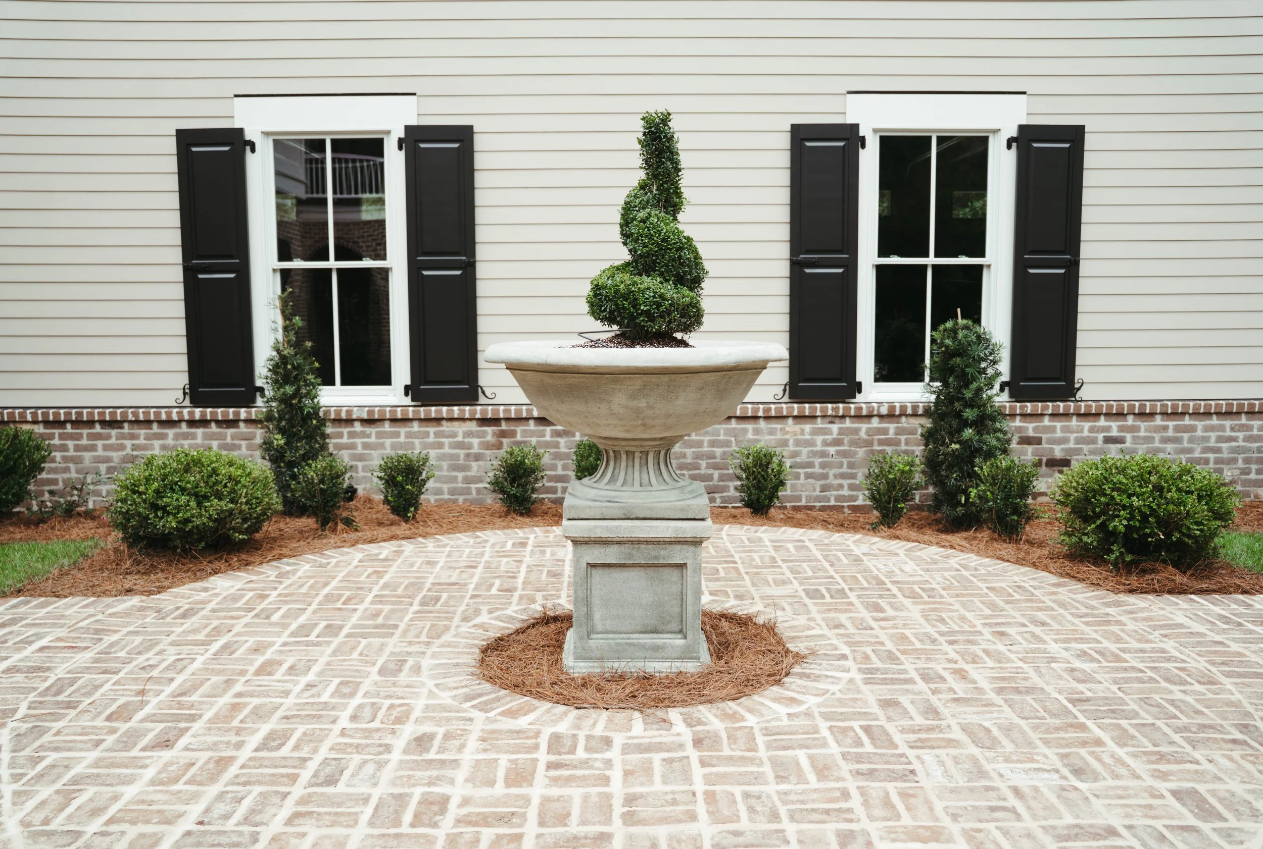 A decorative front yard with a stone planter containing a spiral-shaped topiary, surrounded by a circular brick patio, in front of a house with beige siding, white window frames, and black shutters.