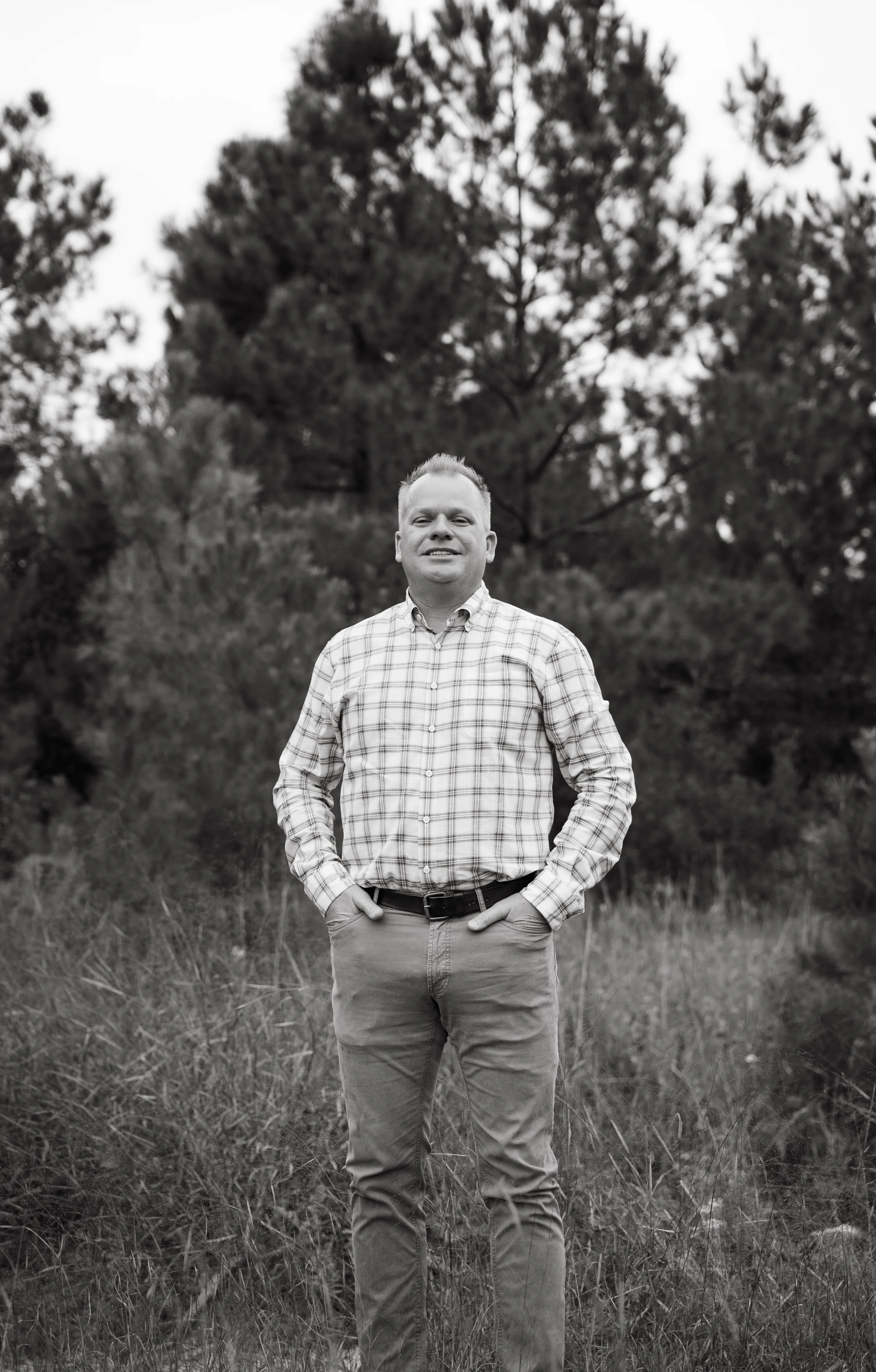 A black and white photo of a man standing outdoors in a grassy area with trees in the background. He is wearing a checkered shirt and light-colored pants, with his hands in his pockets, smiling at the camera.