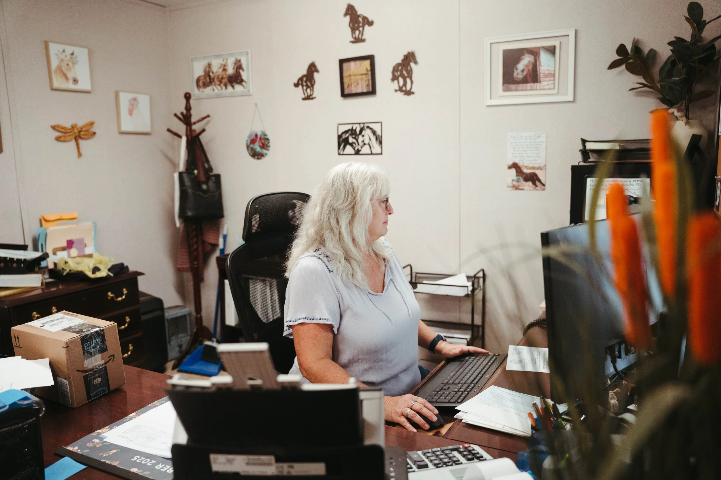 A woman with white hair working at a desk in a home office, surrounded by art and decorations on the wall.