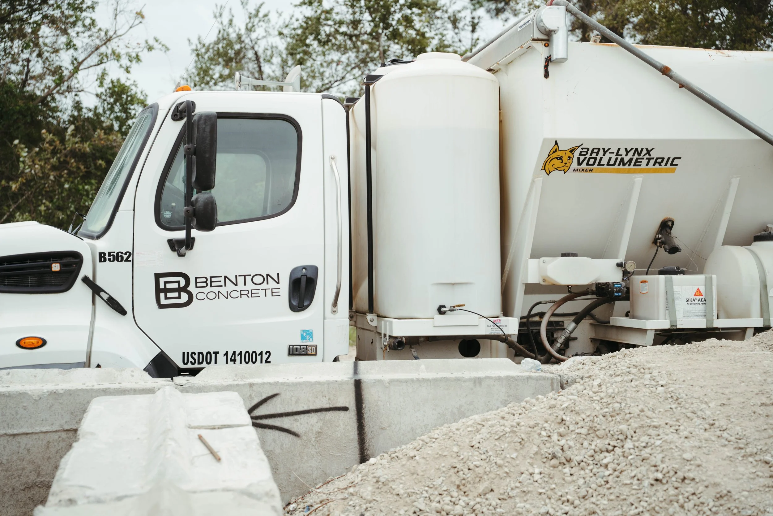 A white cement mixer truck with black and yellow branding, including the logos for Benton Concrete and Bay-Lynx Volumetric Mixer, at a construction site with gravel and concrete blocks in the foreground and trees in the background.