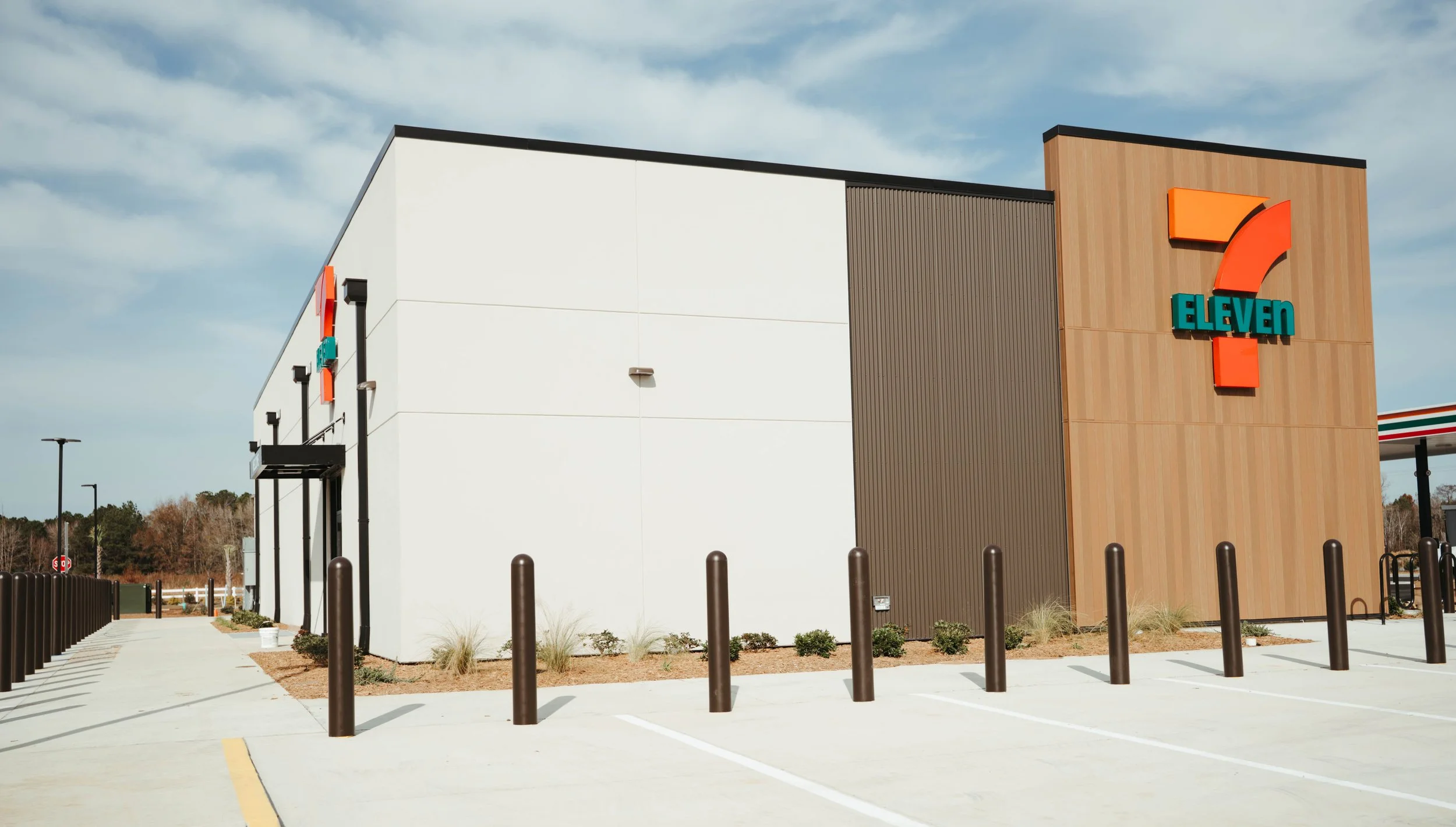 Exterior view of a 7-Eleven convenience store with a large sign displaying the 7-Eleven logo on a wood panel wall, parking spaces, and a partly cloudy sky.