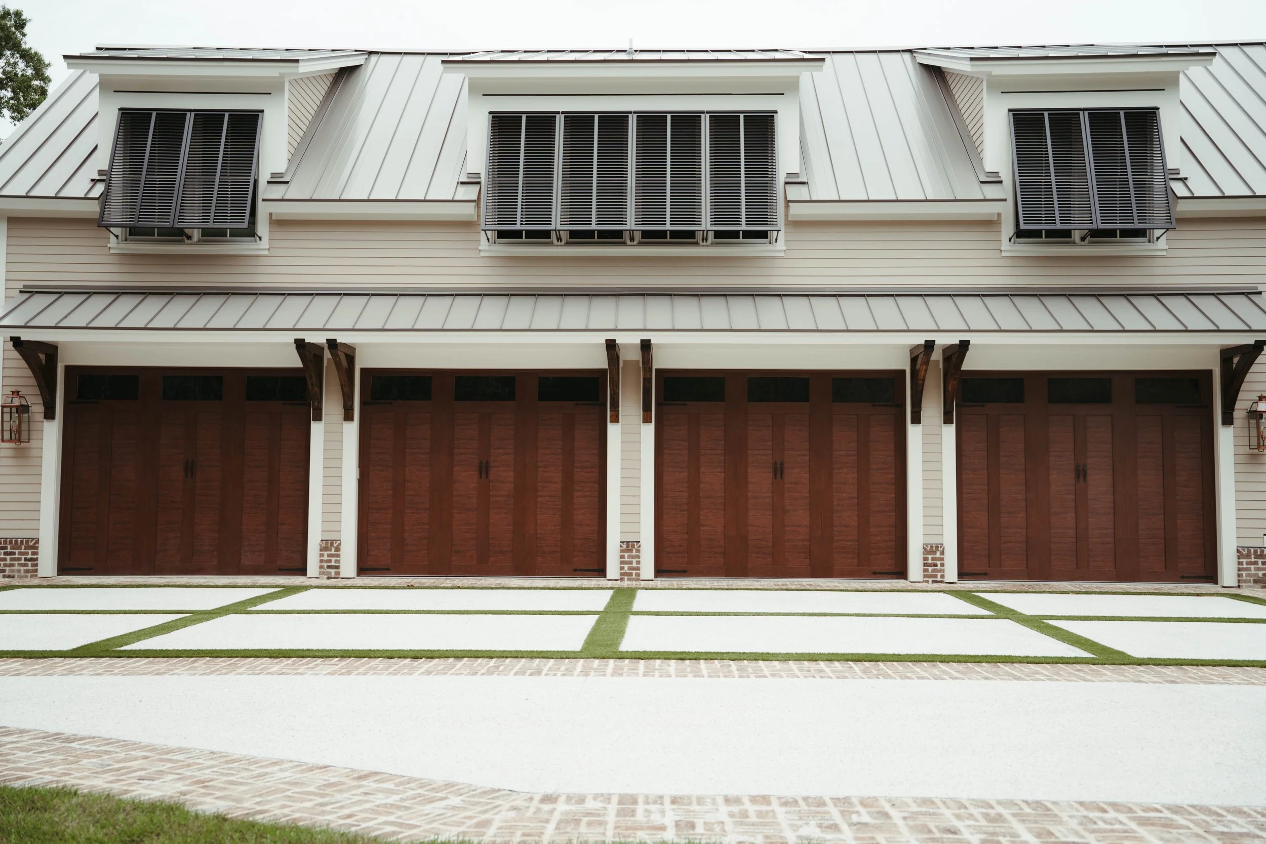 A row of four garage doors on a modern house with stone and white siding exterior, a metal roof, and black window shutters above each garage. The driveway has concrete and brick borders with green grass areas.