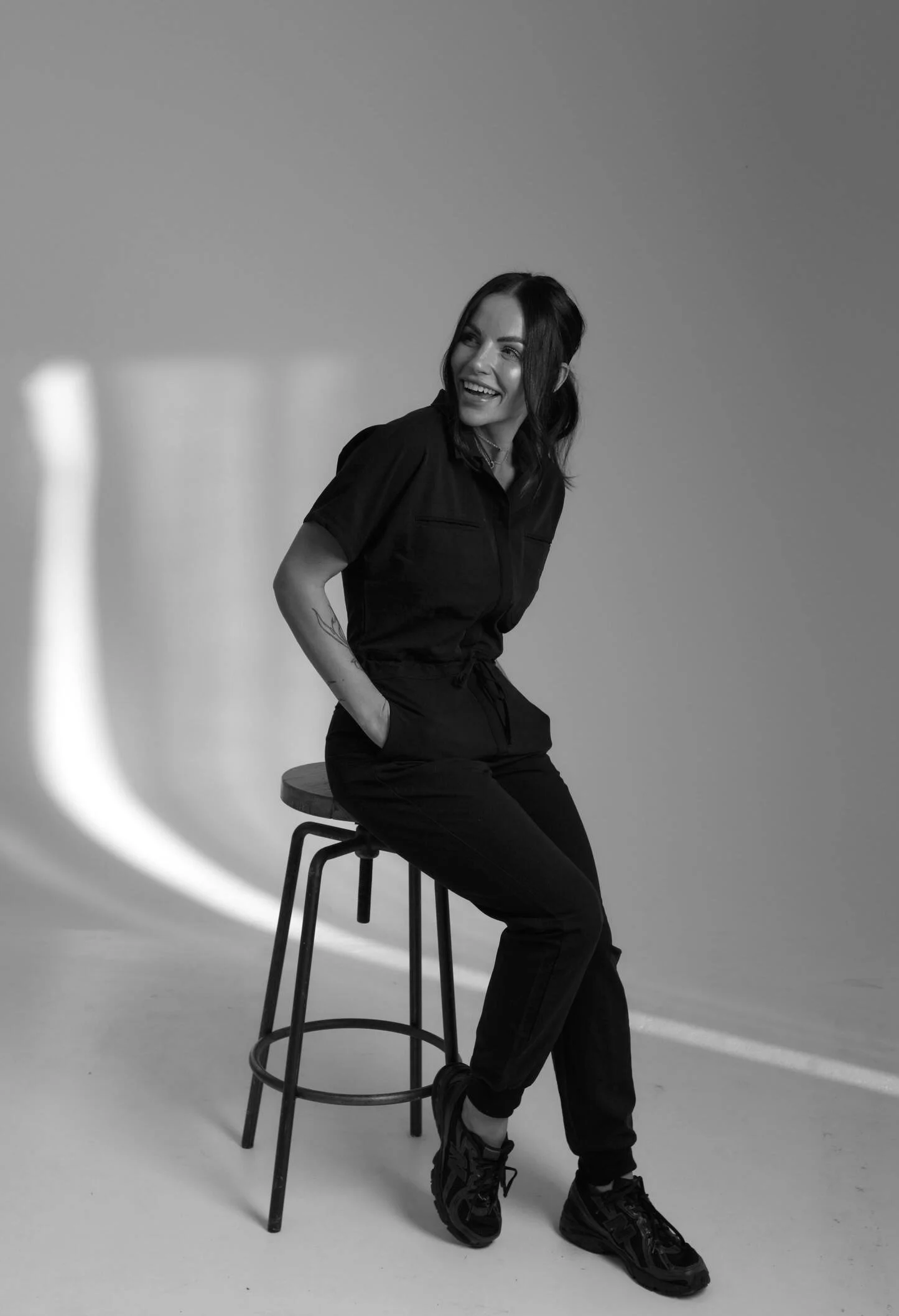 A woman with dark hair is sitting on a stool, smiling, wearing a dark jumpsuit and sneakers, in a studio setting with a plain background and curved lighting.