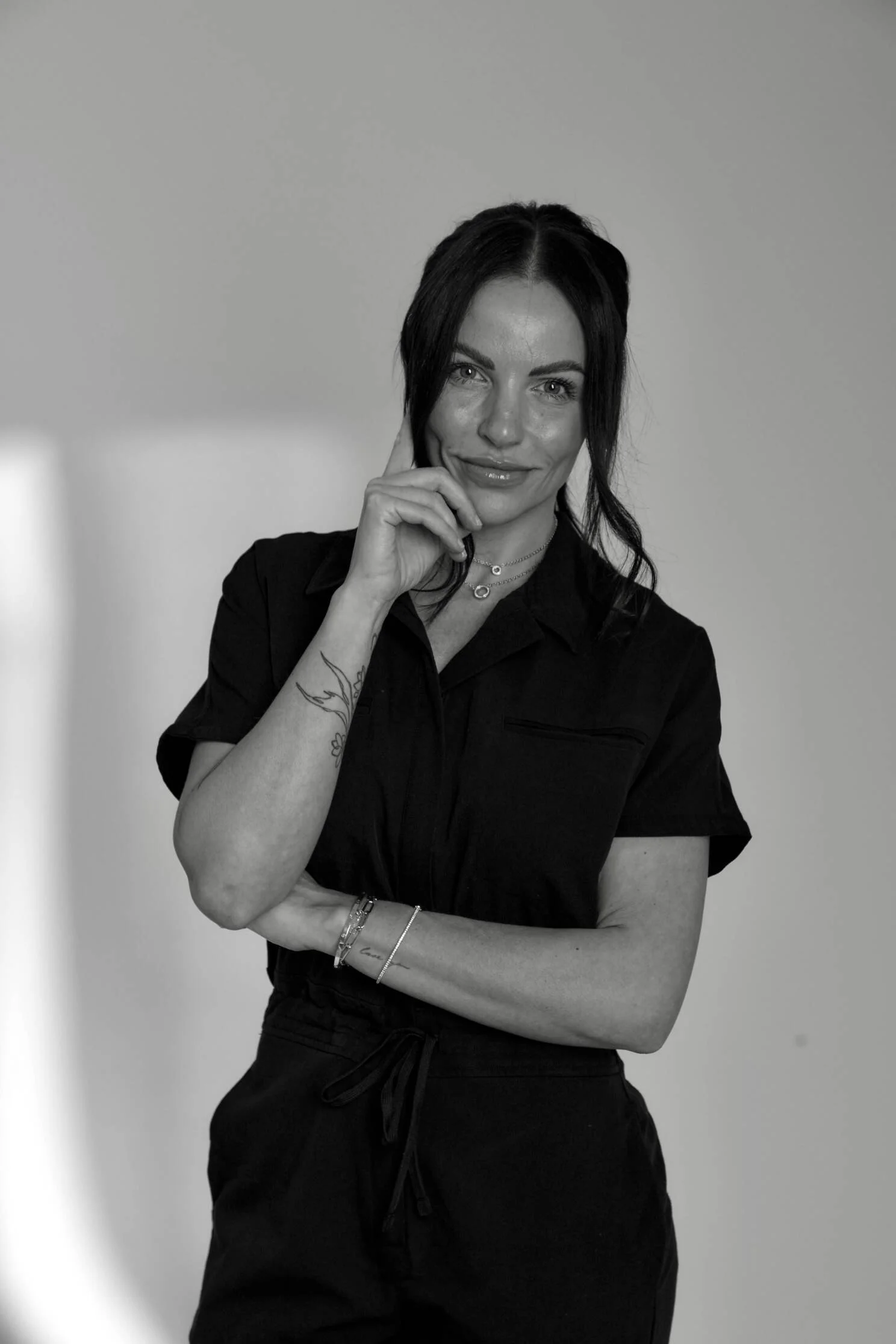 A woman with dark hair, smiling, wearing a dark shirt, posed with her right hand touching her chin, standing against a plain background.
