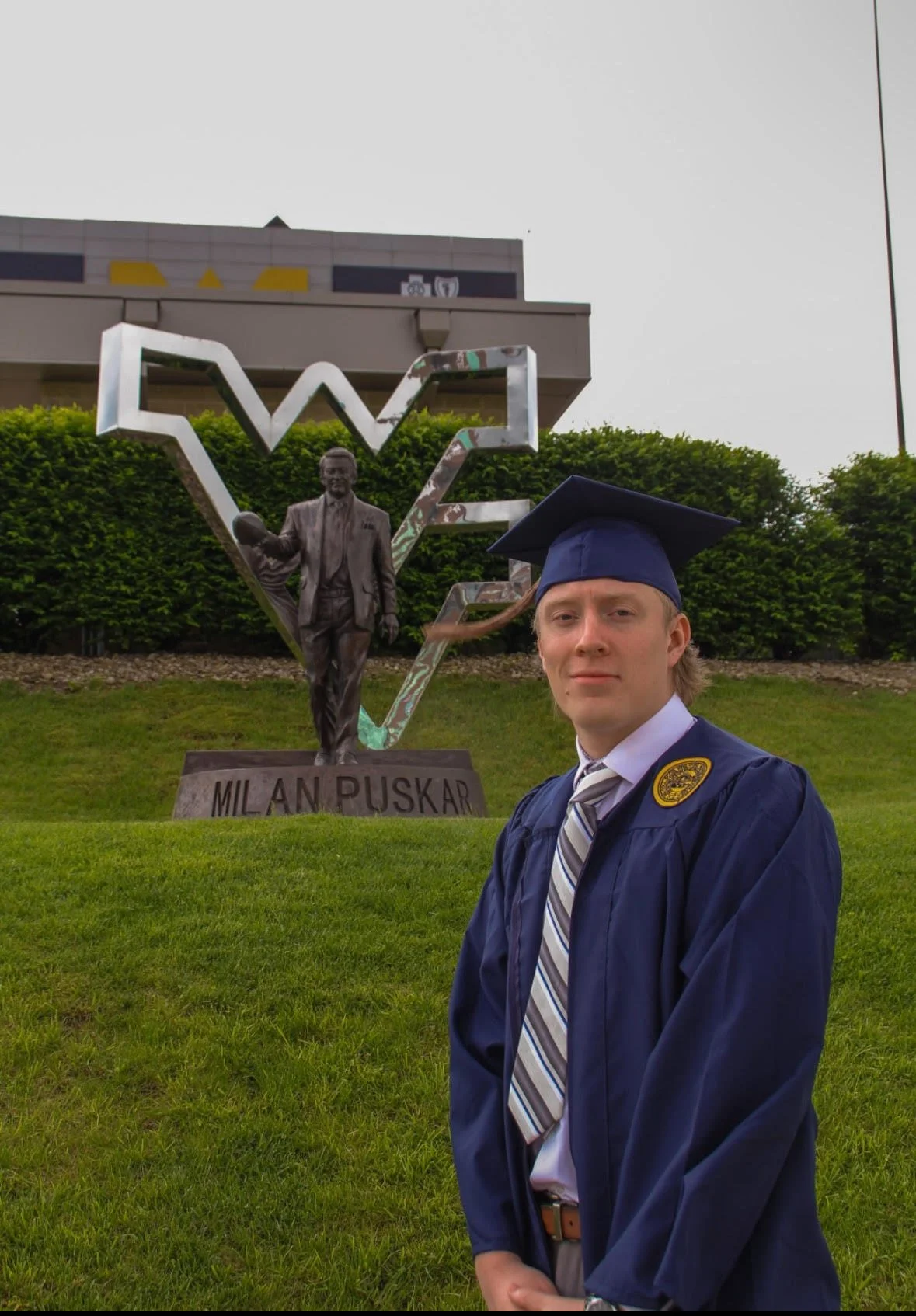 A young man in a graduation gown and cap stands in front of a statue of Milán Rúskar, with a large metallic 'V' and a statue of a man holding a football in the background.
