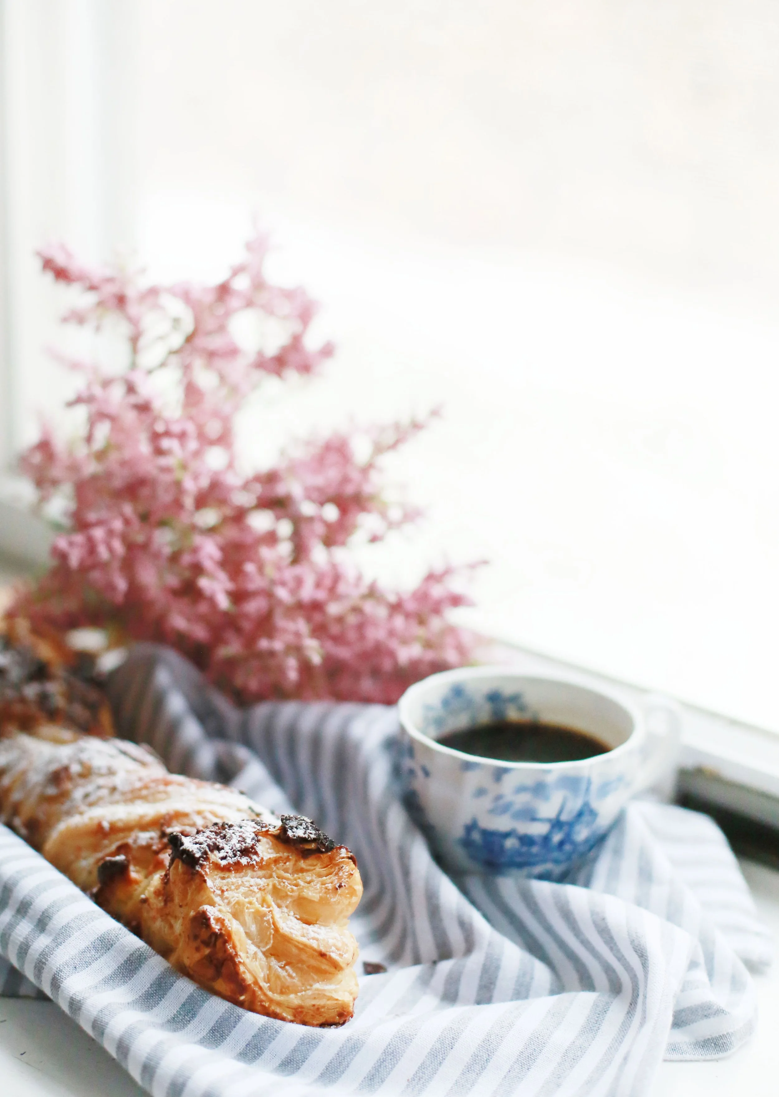 A croissant with chocolate chips on a striped cloth, a cup of black coffee, pink flowers, and a window with bright light.