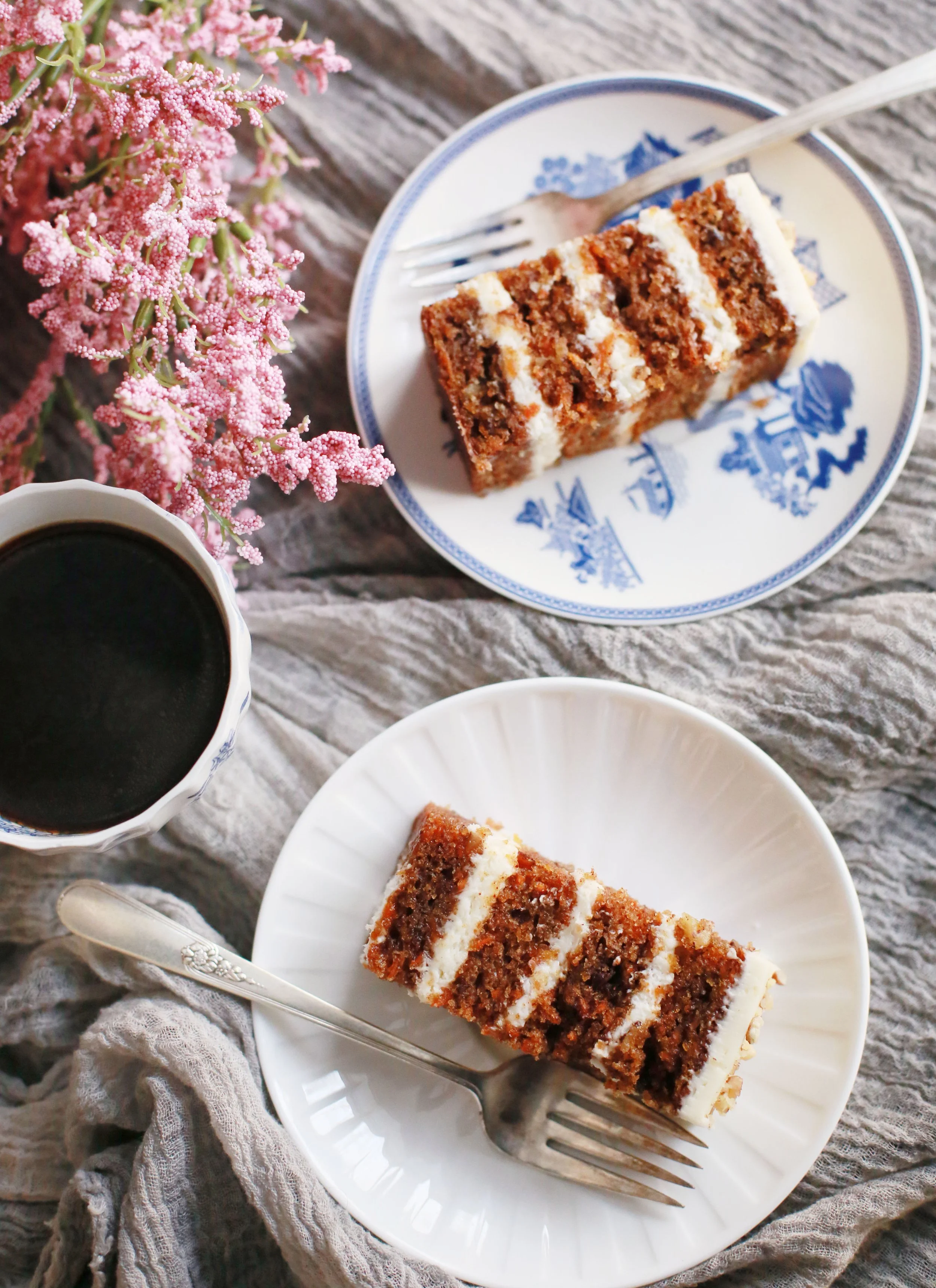 Slices of carrot cake with cream cheese frosting on white and blue plates, a cup of black coffee, pink flowers, and a gray textured cloth on a rustic wooden surface.
