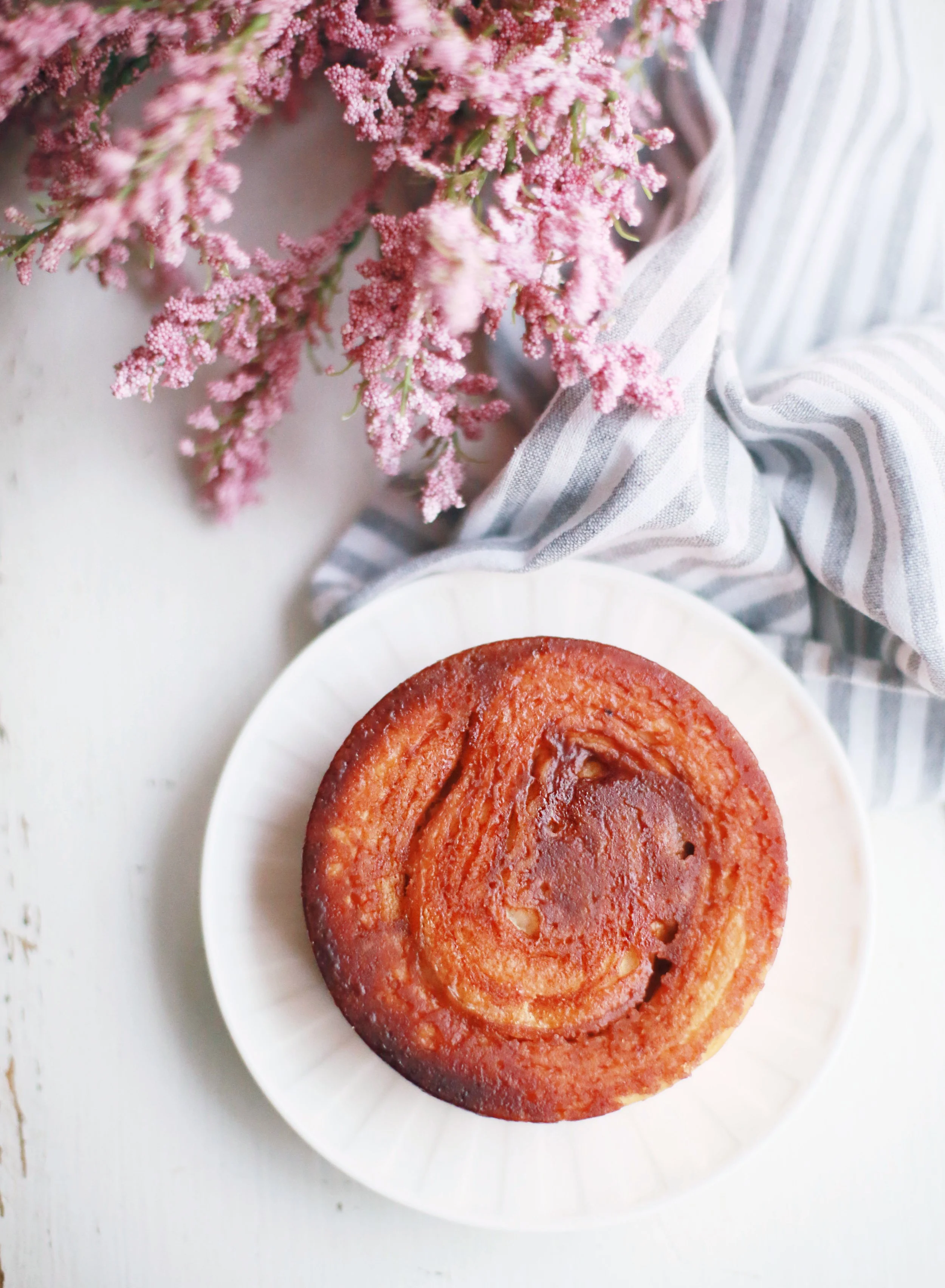 A vanilla or yellow cake with a glazed or caramelized top on a white paper plate, with pink flowers and a striped cloth nearby.