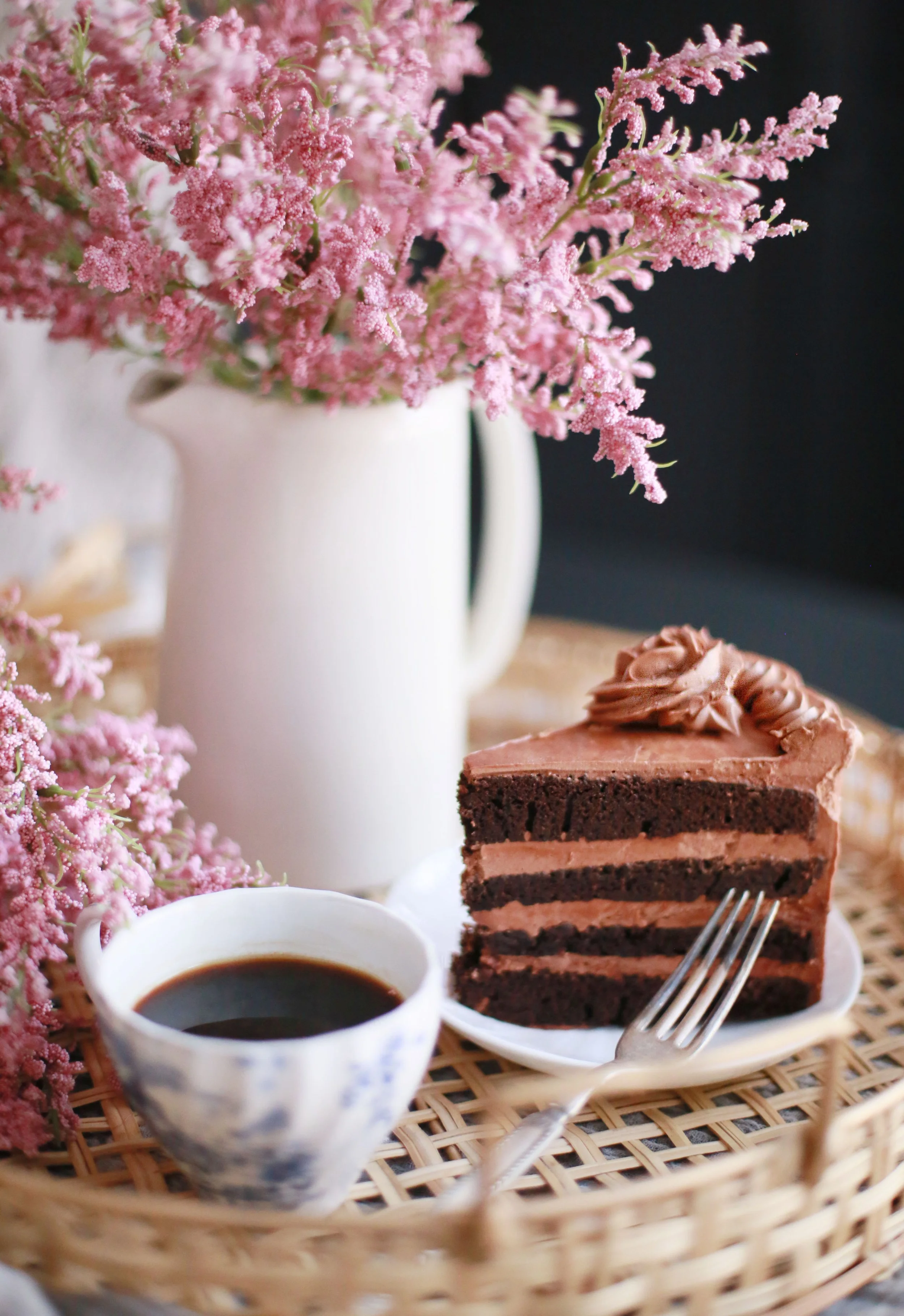 A slice of chocolate cake with pink frosting on top, a cup of black coffee, a white pitcher filled with pink flowers, all arranged on a woven tray.