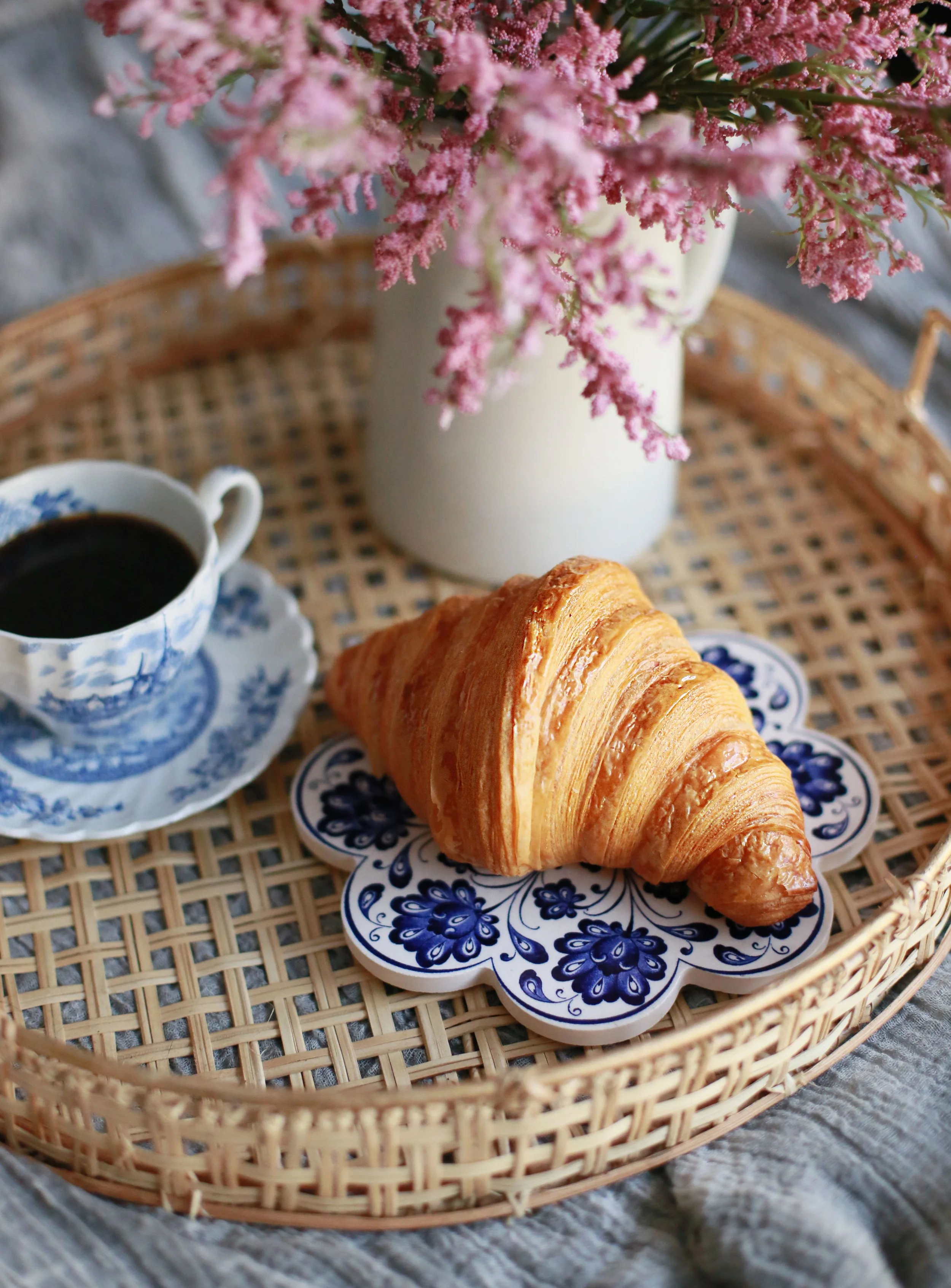 A croissant on a blue floral plate, a cup of black coffee in a blue and white cup and saucer, and a bouquet of pink flowers in a white vase on a wicker tray.