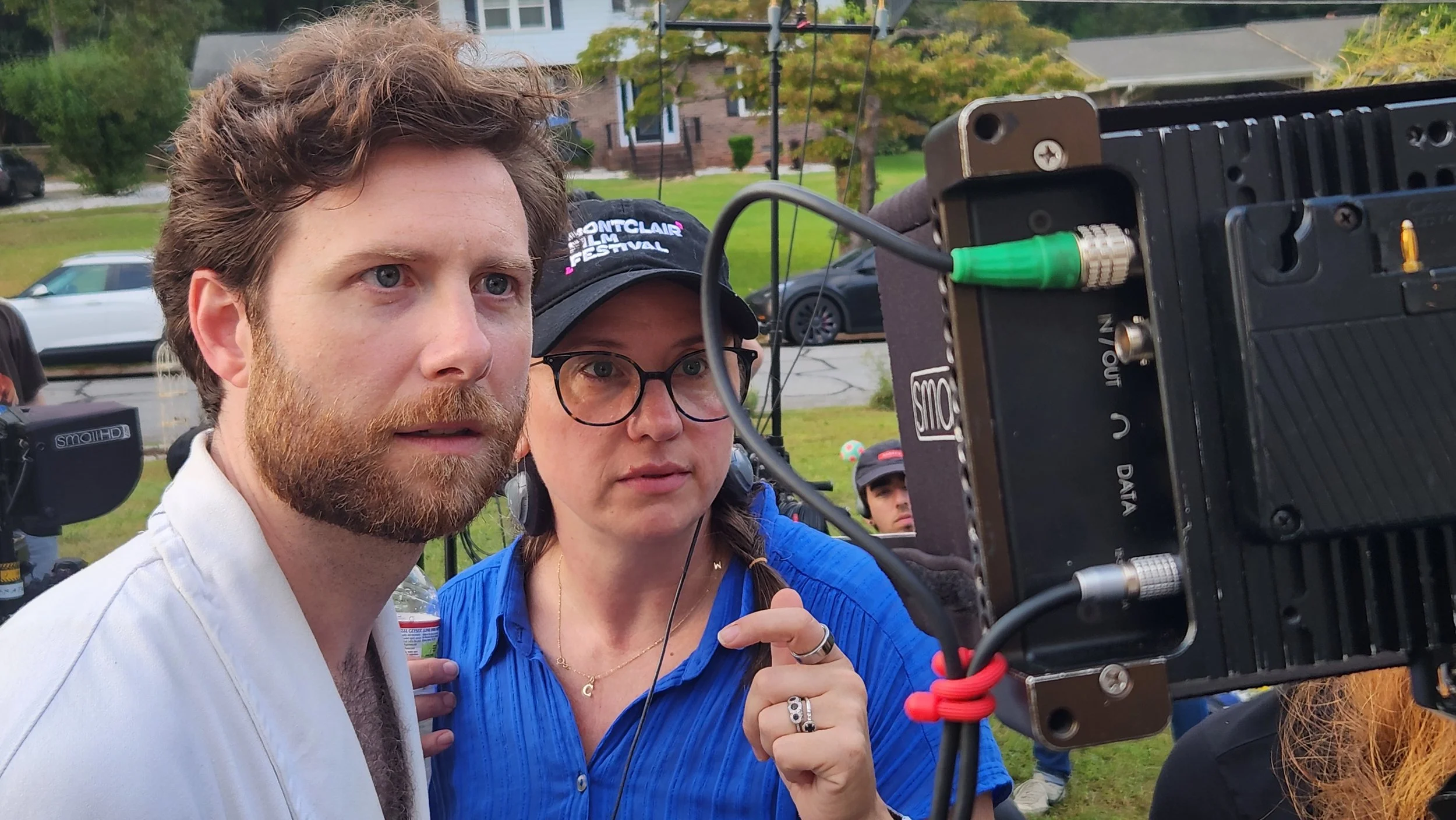 Two people looking at a camera monitor outdoors, with a man having brown hair and a beard, and a woman with glasses and a baseball cap that says "Montclair Film Festival."