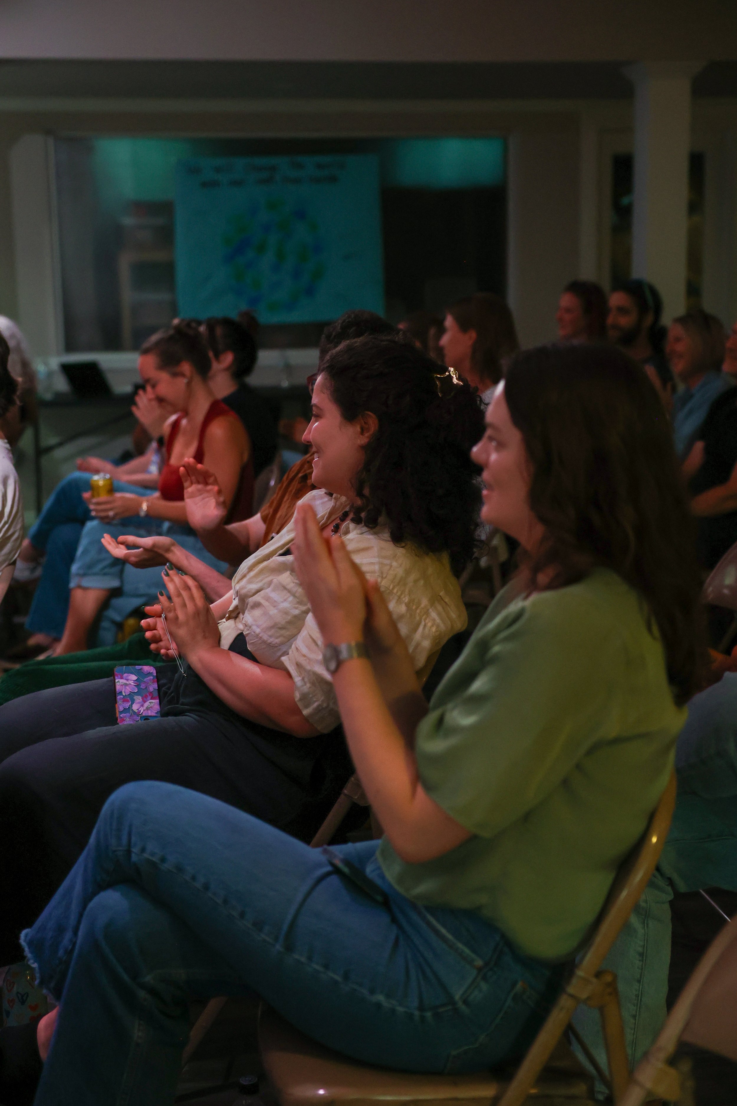 People sitting in an audience, smiling and clapping, at a presentation or event in an indoor setting.