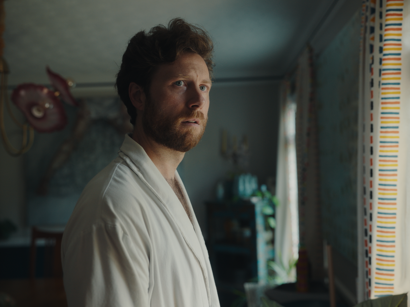 Mr Jesus's main character, Chris, a man with a beard and curly hair looking toward a window with curtains in a room.