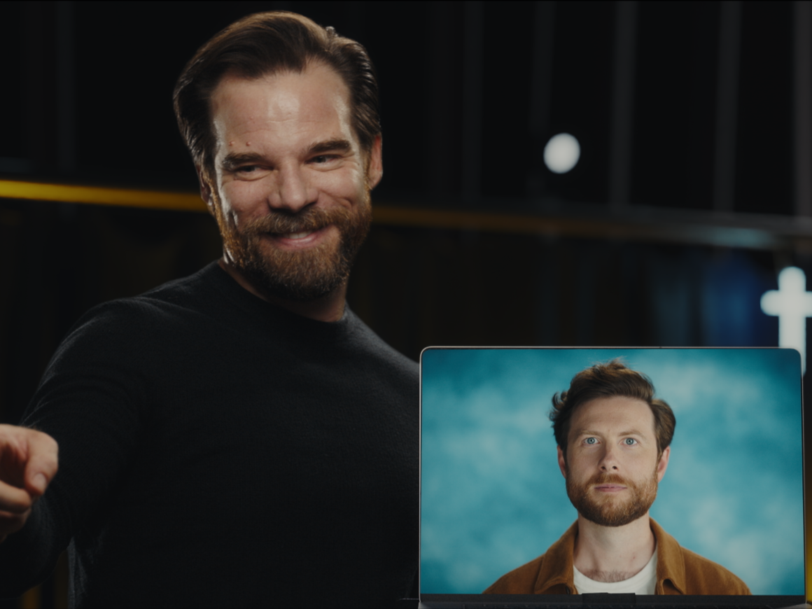 A man with a beard smiling at a camera, holding a laptop with a video call showing another man with a beard and light brown hair against a blue background.