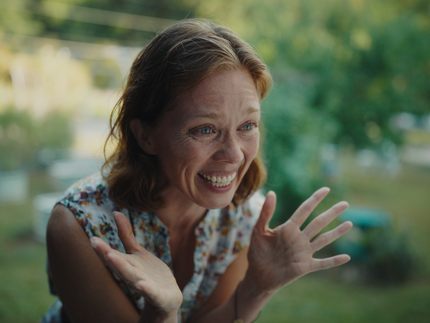 A woman with reddish-brown hair and blue eyes smiling happily outside, wearing a sleeveless floral top, with a blurred background of green trees and sunlight.
