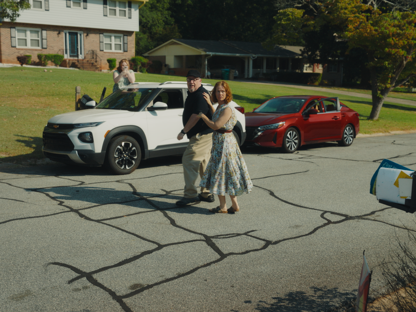 A man and a woman holding hands walking on cracked pavement in a residential area. In the background, a white SUV and a red sedan are parked on the street, with a woman taking a photo nearby. Houses and trees are visible behind them.