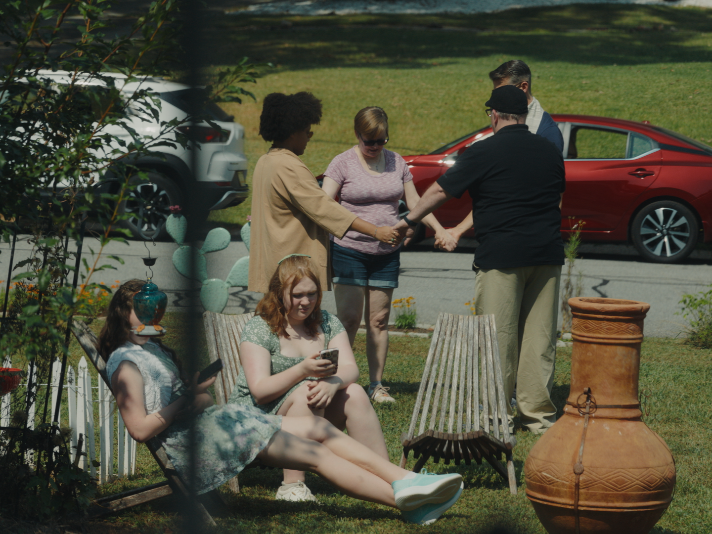 People gathered in a backyard, holding hands in a circle during a prayer or moment of silence, with two women sitting on lawn chairs using their phones.