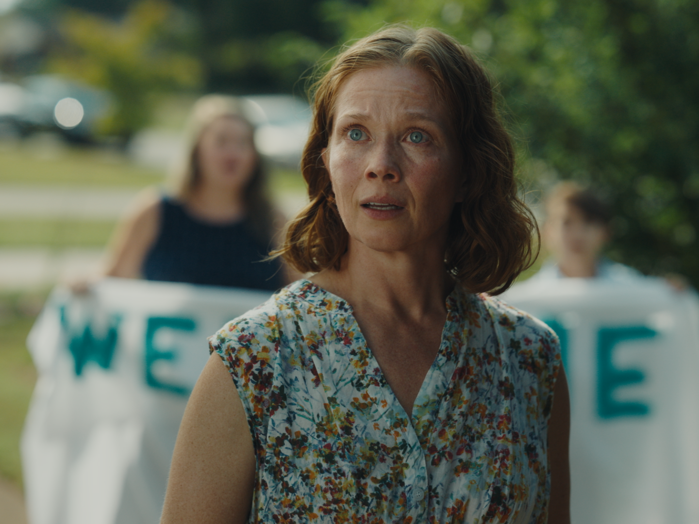 A woman with short curly red hair and blue eyes looks concerned, wearing a floral sleeveless dress, with women holding signs in the background, outdoors with trees and cars.
