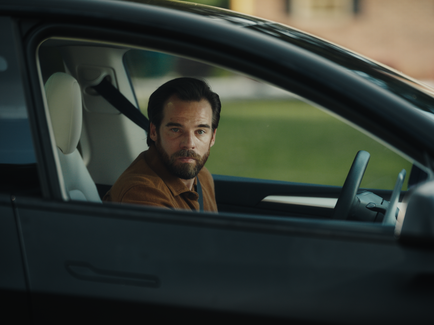 A man with dark hair and a beard sitting in the driver's seat of a car, looking out through the window with a serious expression.
