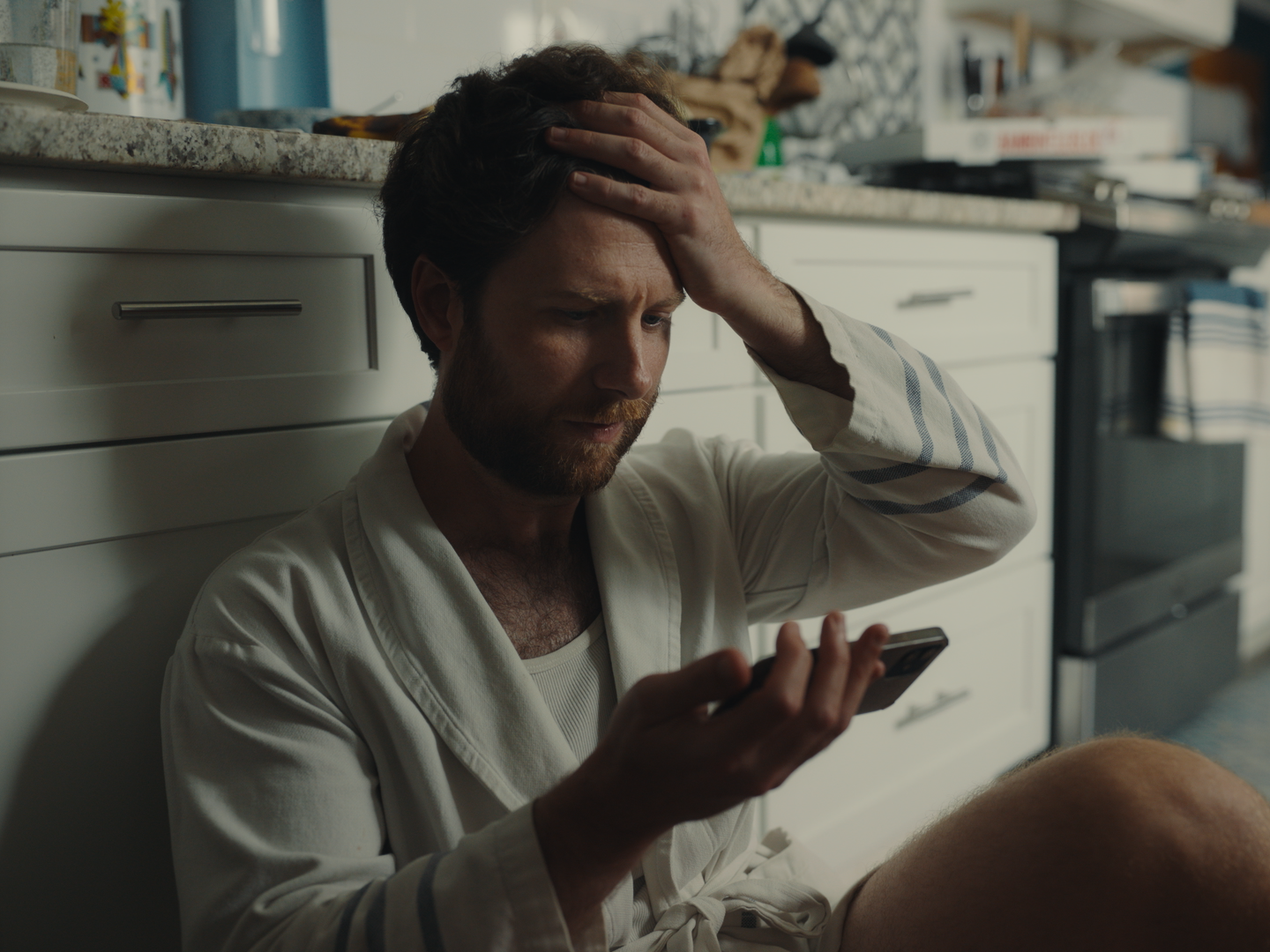 A man with a beard and curly hair sitting on the kitchen floor, holding his head and looking at his phone with a frustrated or worried expression.