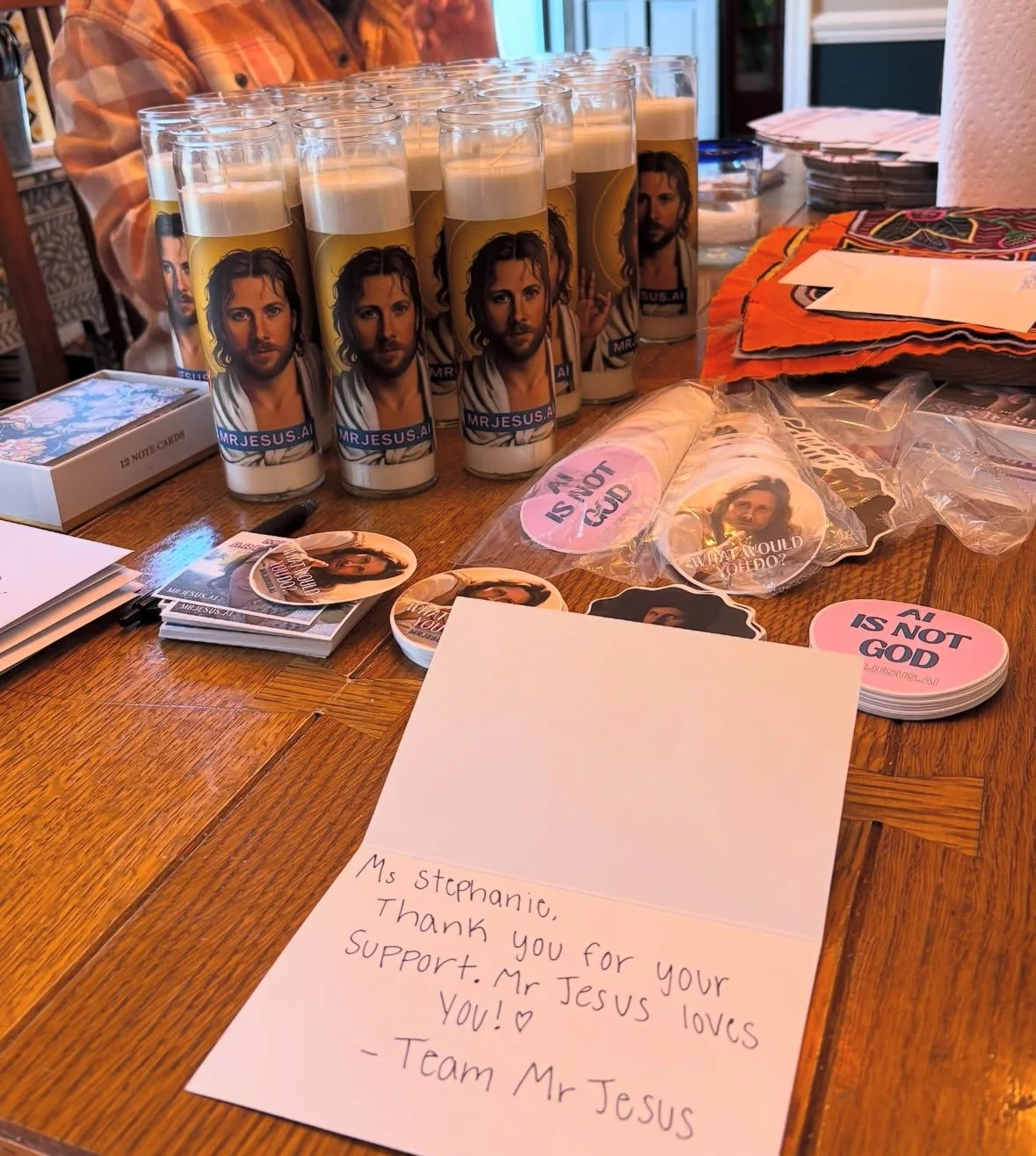 Table with religious-themed items including candles featuring an image of Jesus, circular stickers with a Jesus image and the message 'AM I NOT GOD,' notecards with a handwritten note, pens, and cloth napkins.