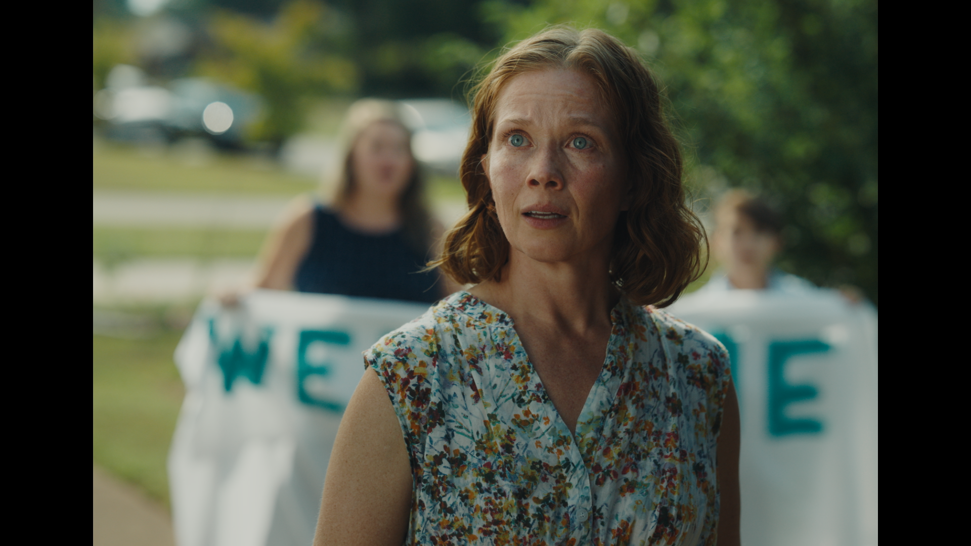 One of the film's main characters, Anna, a woman with red hair and blue eyes standing outdoors, with a woman and two small children holding Welcome Back signs in the background.