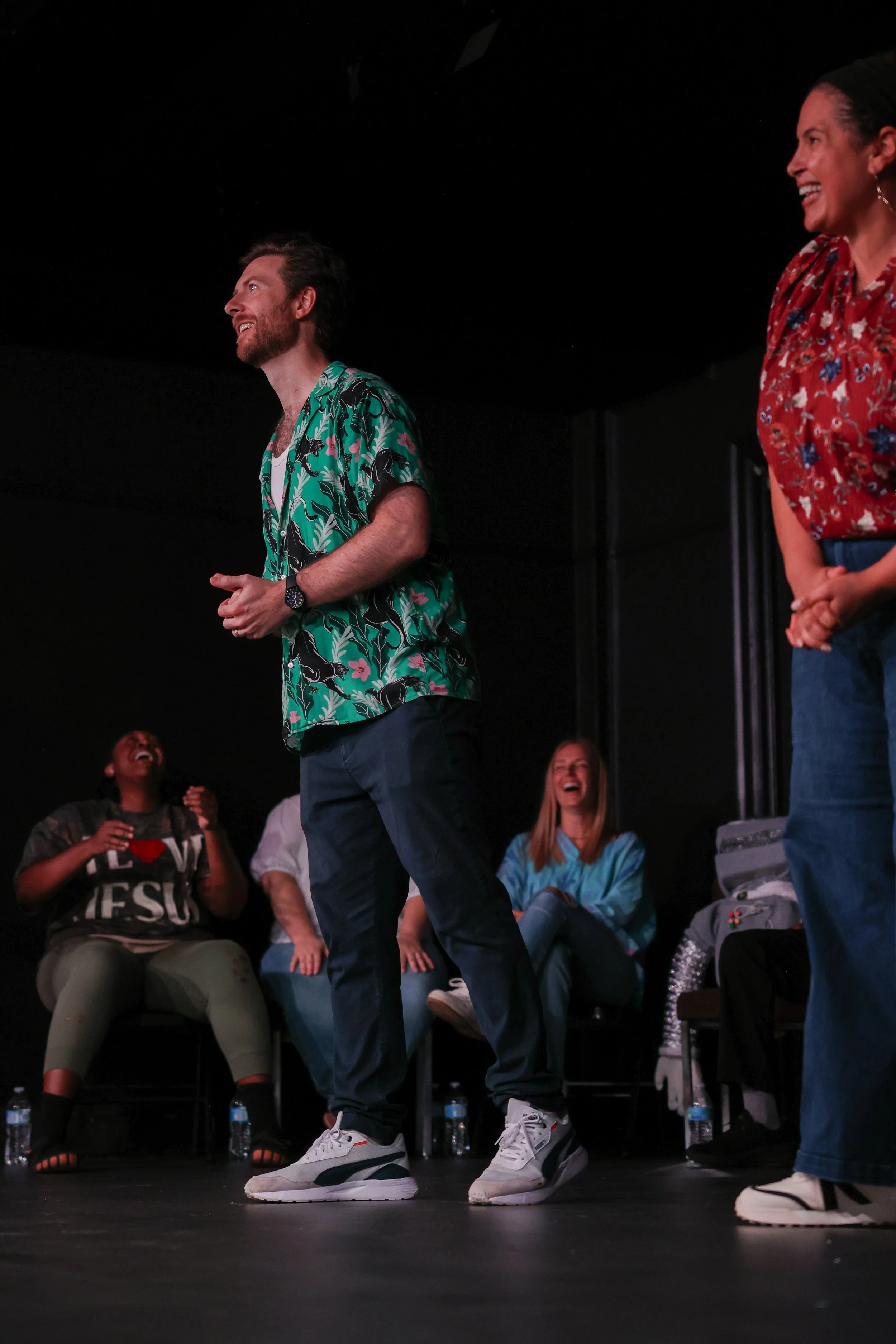 Group of people laughing on stage, with two individuals standing in the foreground and others seated in the background.