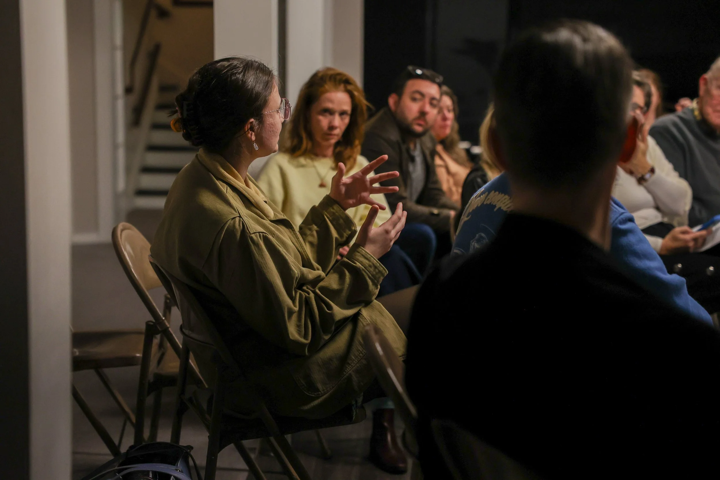 A woman with glasses and a jacket is speaking and gesturing with her hands during a discussion or meeting, surrounded by several seated people in a dimly lit room.
