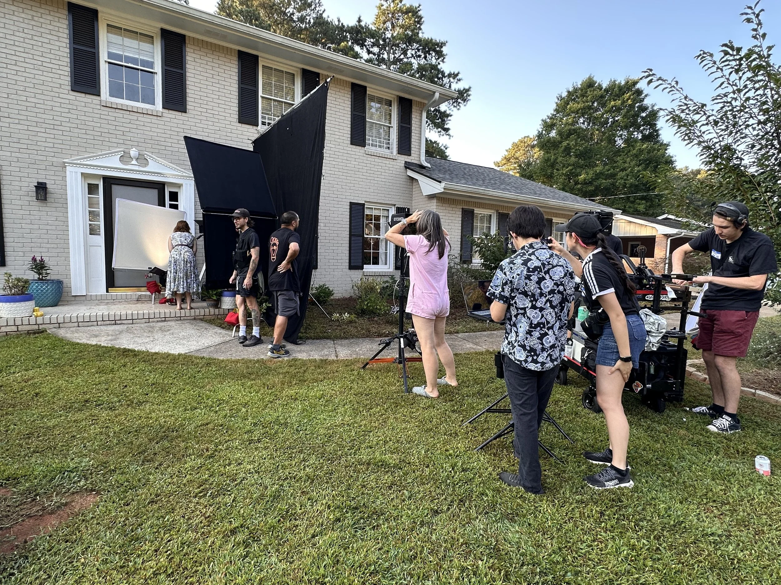 Filming crew sets up equipment outside a house as actors prepare for a scene.