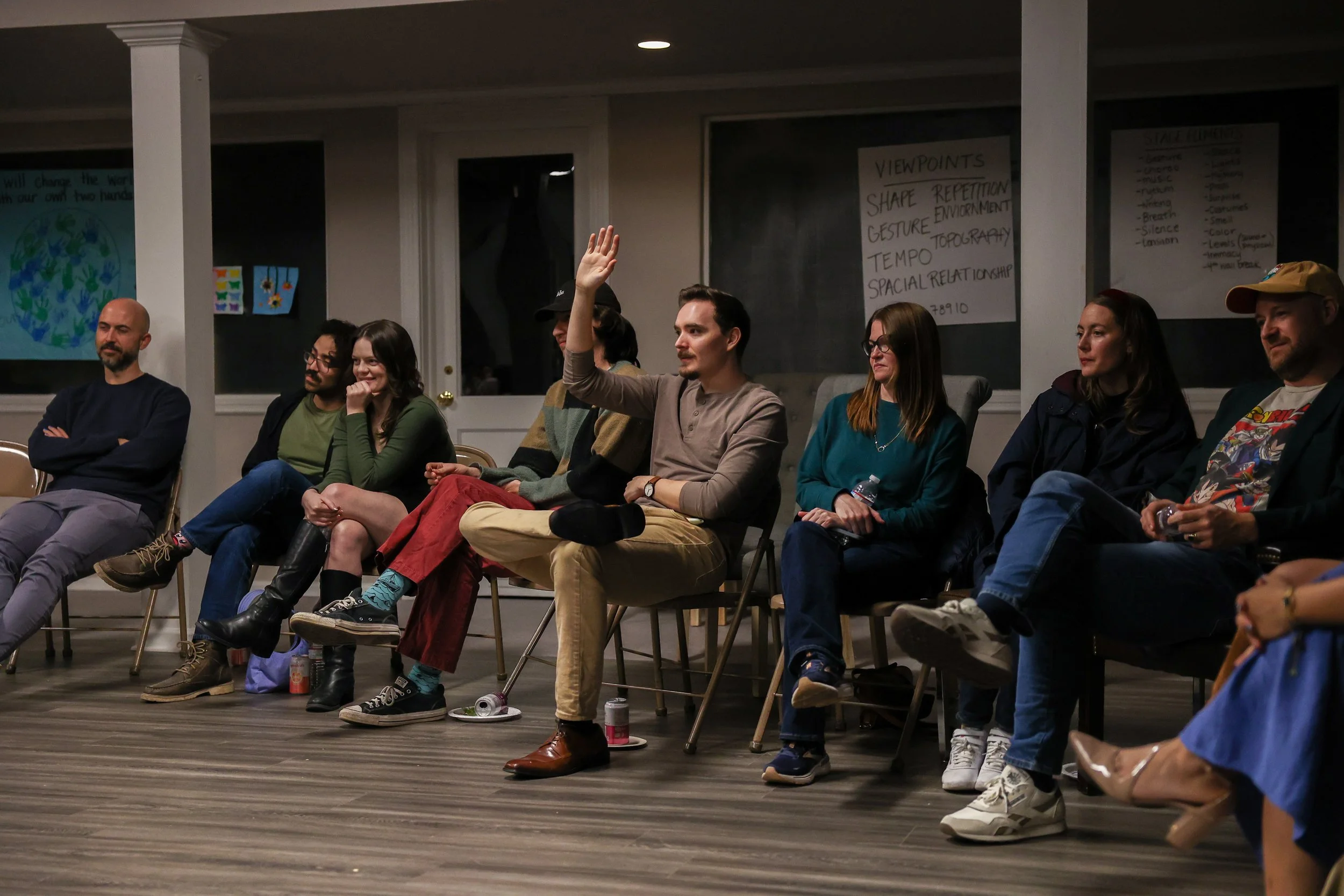 A group of people sitting in a semicircle in a room, one man in the middle raising his hand. The room has posters and notes on the walls, and the floor is wooden.