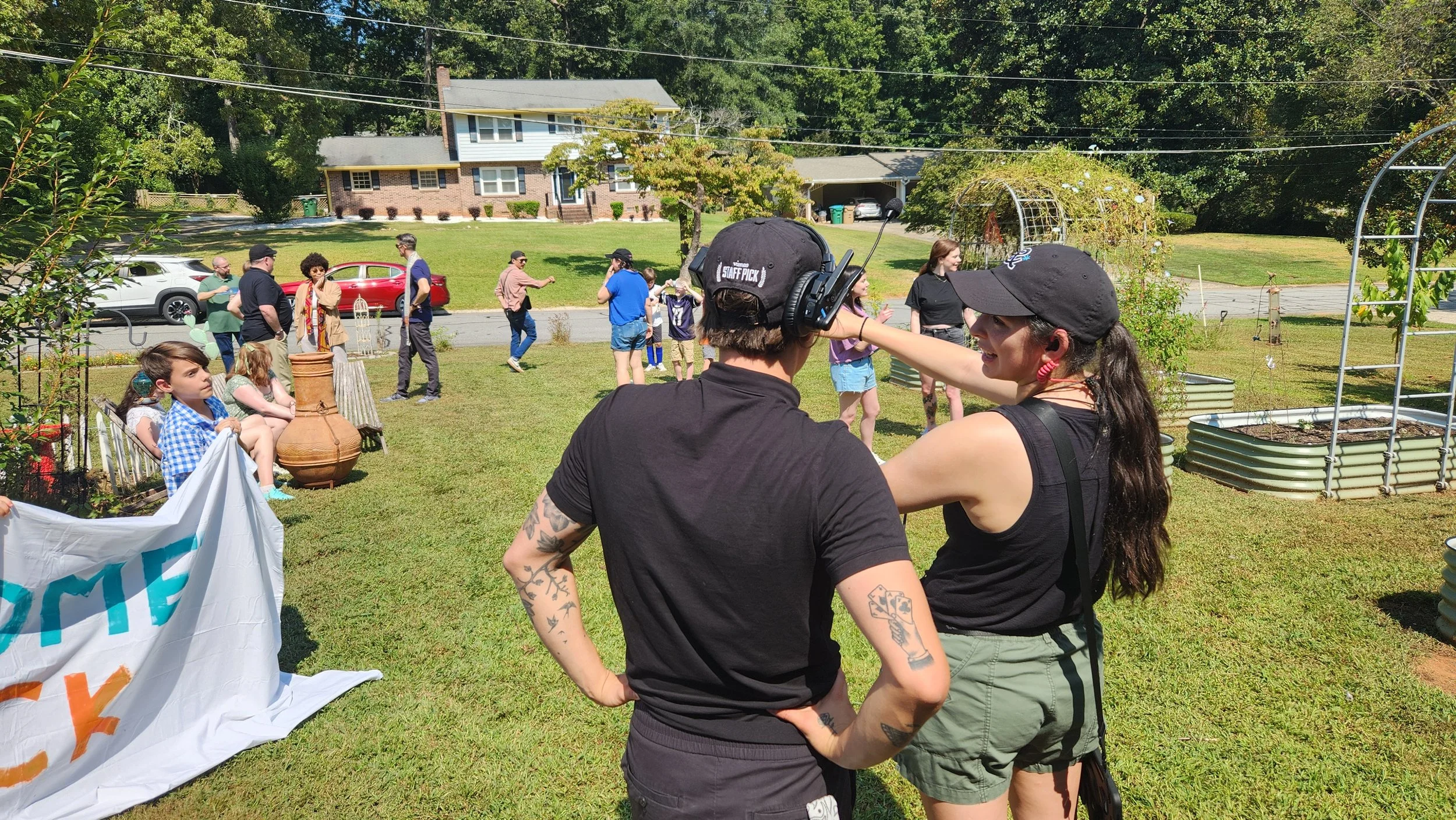 Two people in the foreground, one wearing a black t-shirt, cap, and headphones, and the other wearing a black tank top, cap, and earrings, are engaged in conversation at an outdoor gathering. In the background, children and adults are mingling on a grassy yard with houses and trees, some sitting and others walking or talking. There are garden beds and decorative items nearby.