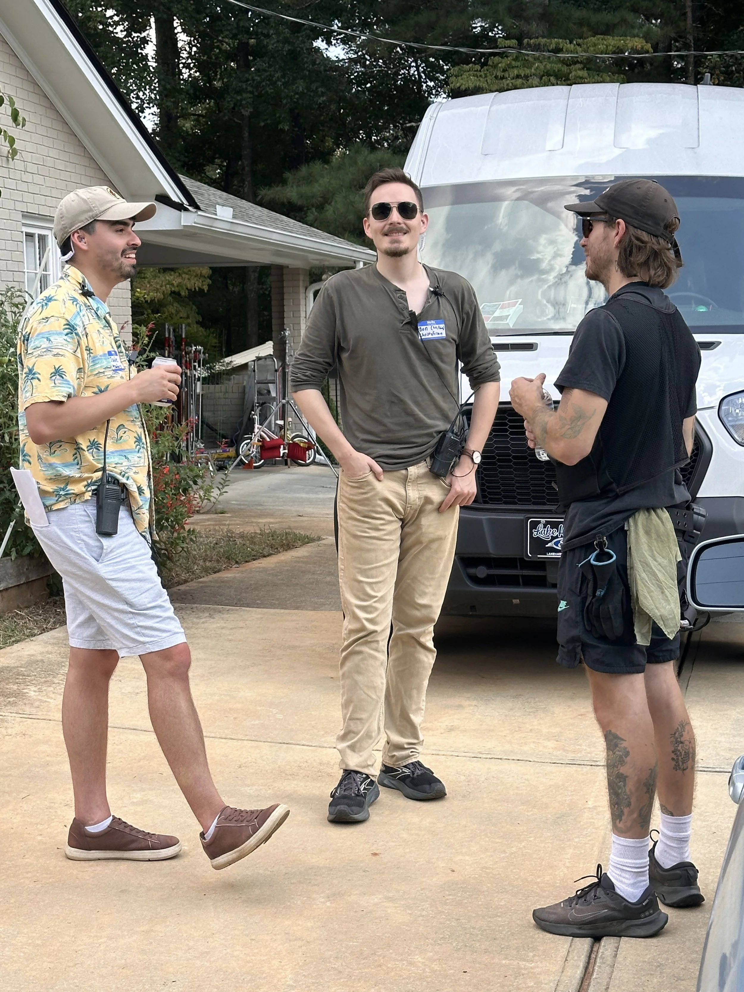 Three men standing outside near a white van, engaged in conversation, with one man smiling and wearing sunglasses, dressed casually, and the other two men also casually dressed with one wearing a Hawaiian shirt and the other with tattoos on his legs.