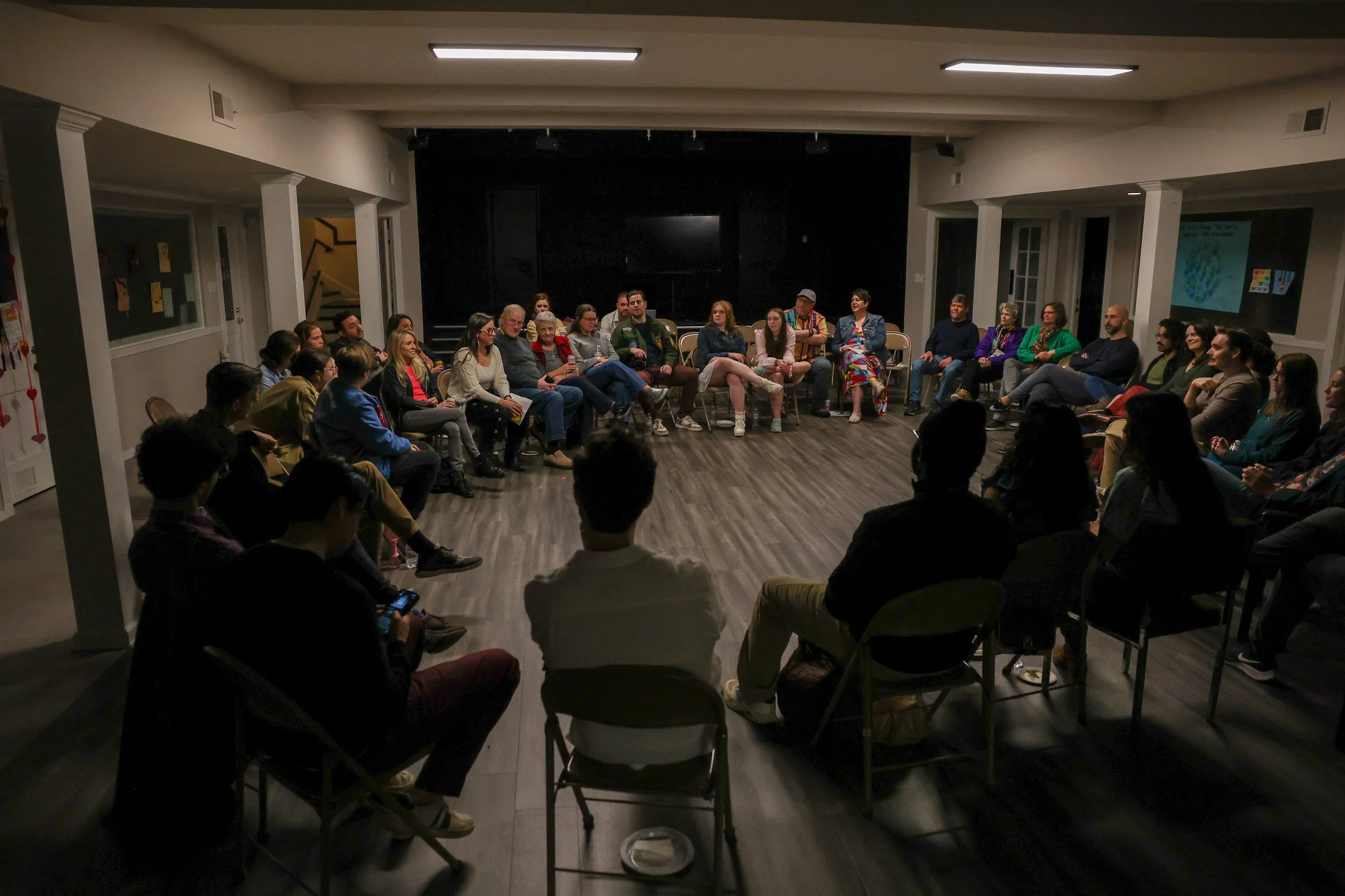 A group of people seated in a large circle in a dimly lit room, participating in a discussion or meeting.