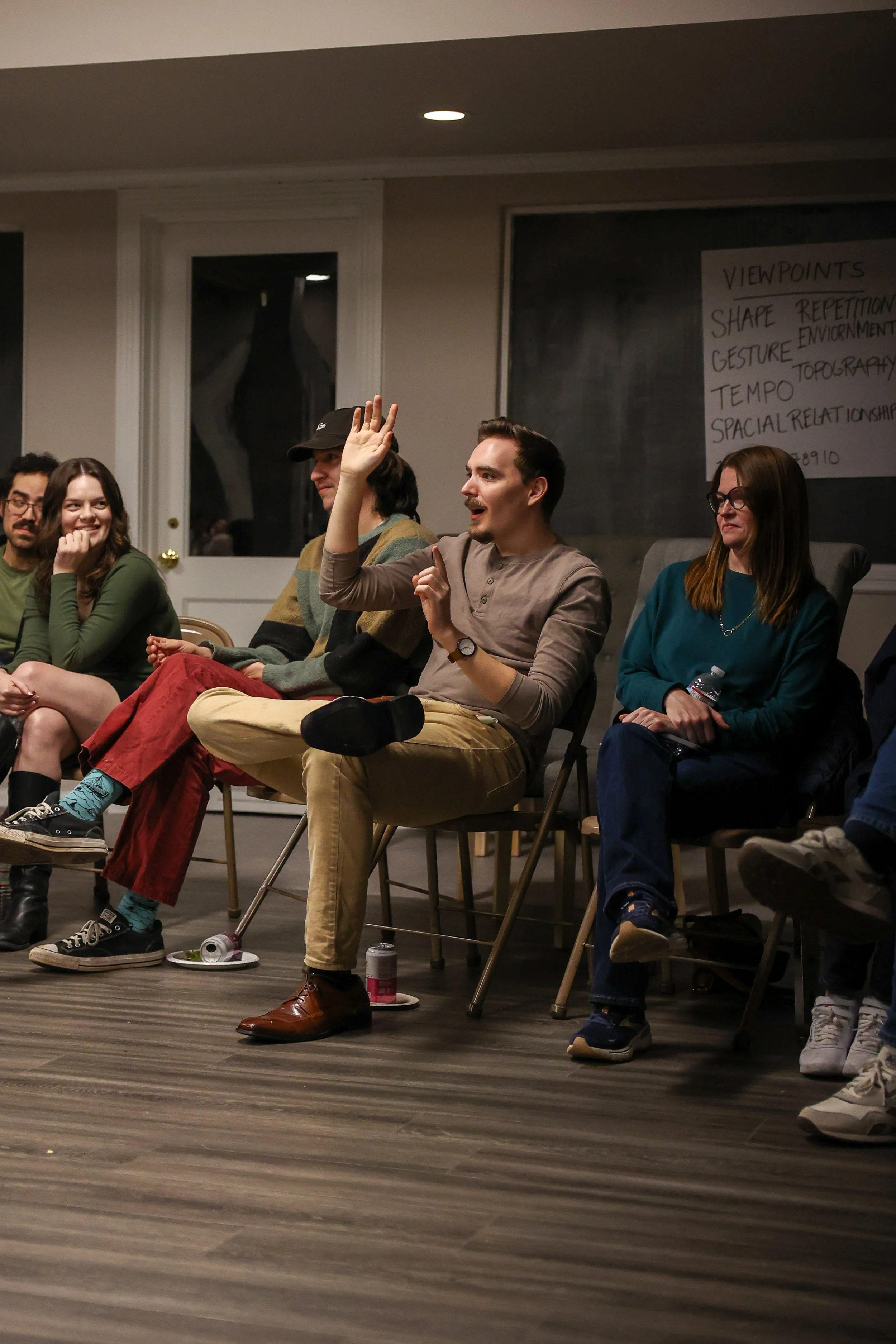 A man in a gray shirt and beige pants speaking and raising his hand in a group discussion or meeting, with four other people seated around him, in a room with a blackboard and a note sheet in the background.