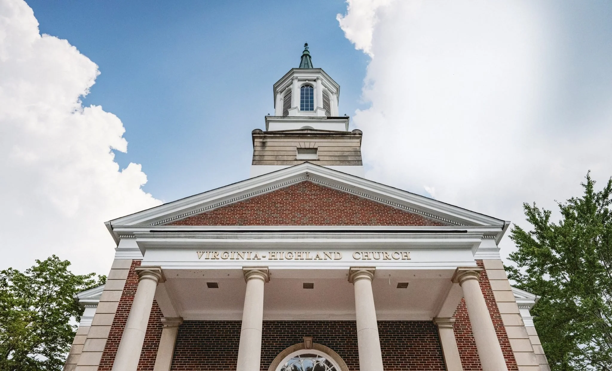 Front view of Virginia Highland Church with brick facade, white columns, triangular pediment, and steeple against a partly cloudy sky, surrounded by green trees.