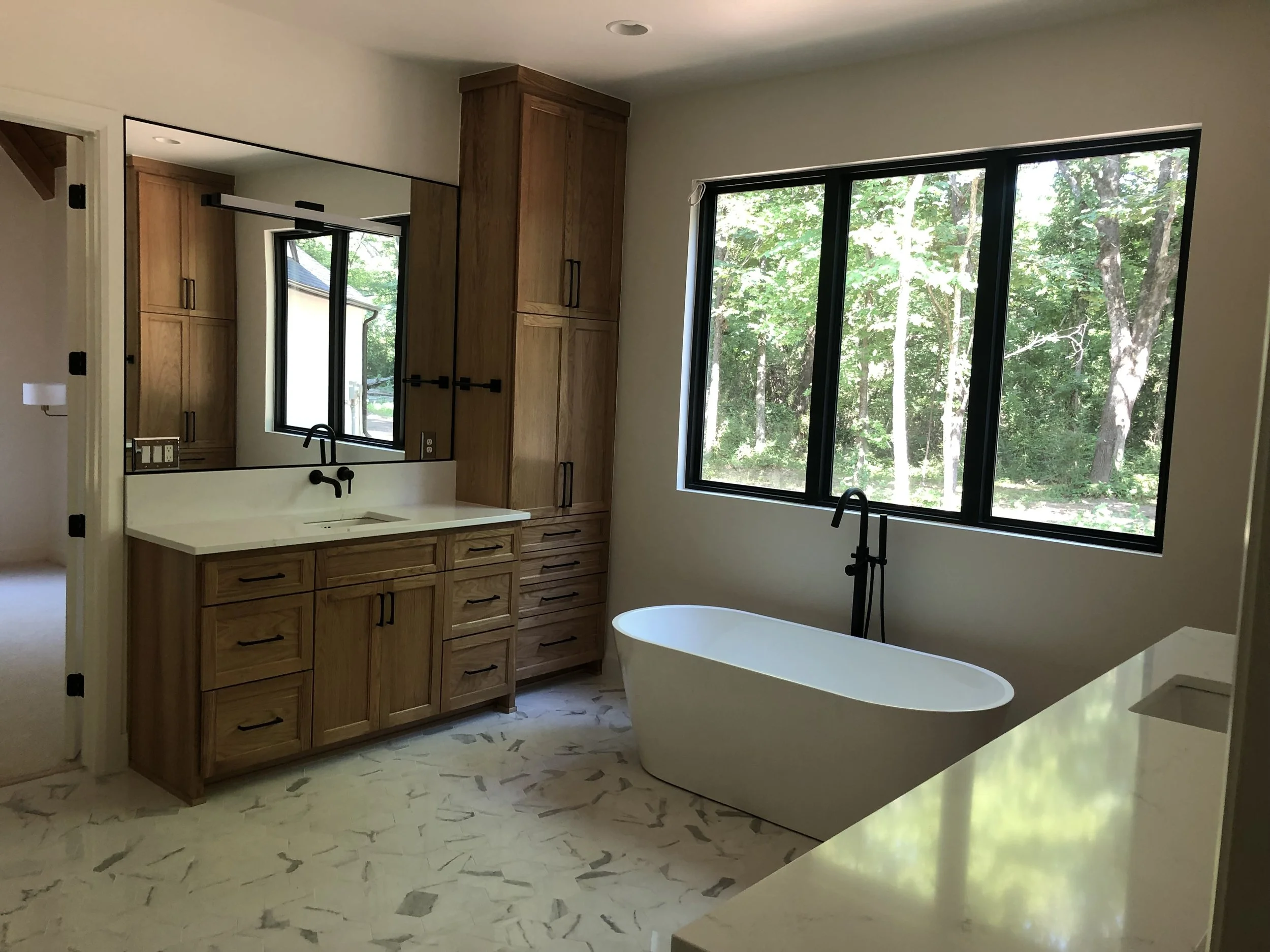 Modern bathroom with a freestanding bathtub beneath large window, wood cabinets, and a large mirror above the sink with black fixtures.
