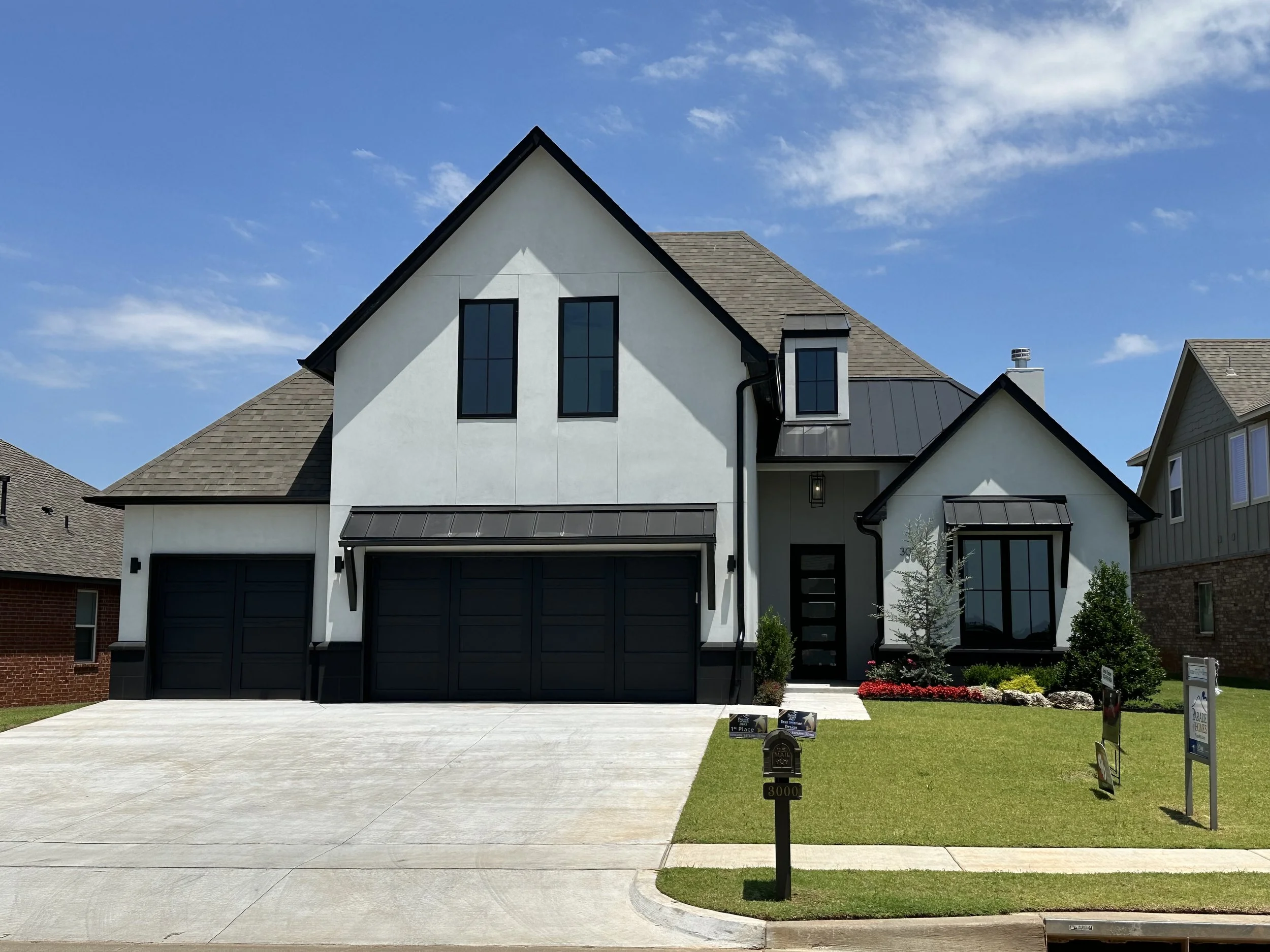 New modern two-story house with white facade, black accents, and a three-car garage, front yard with grass, trees, and mailbox, under a clear blue sky.