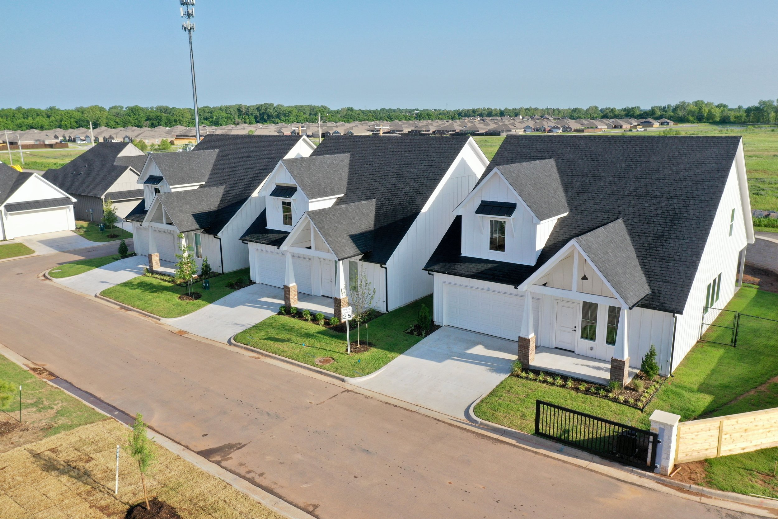 Aerial view of new white modern houses with black roofs in a suburban neighborhood, with green lawns, driveways, and a paved street.